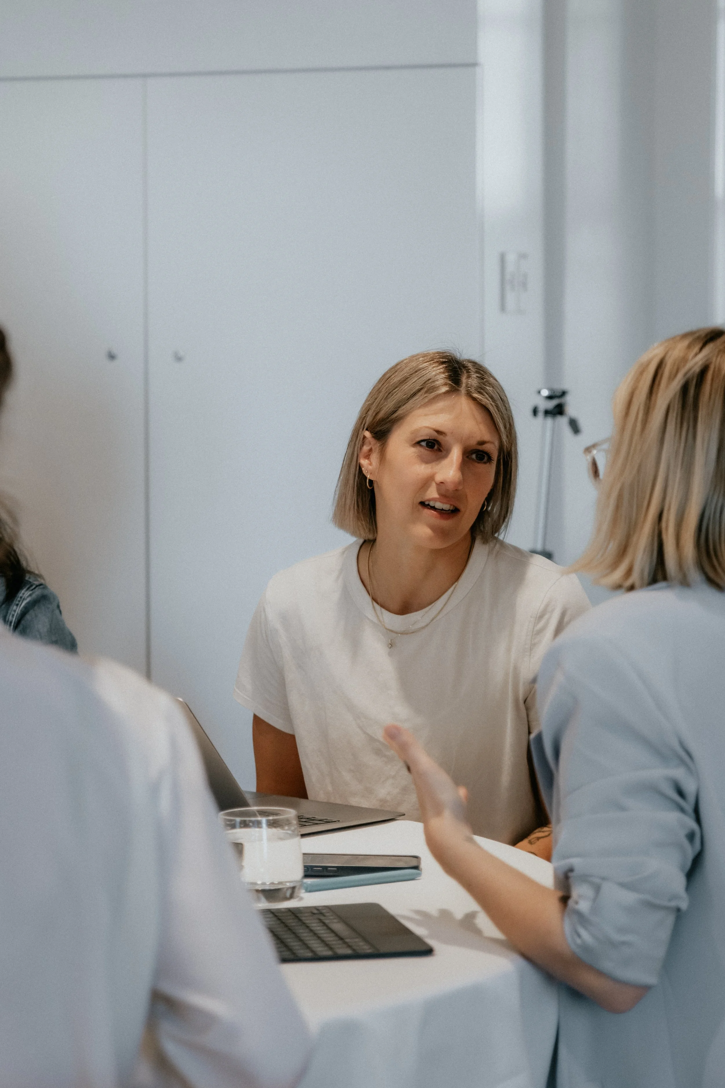 Group of people having a conversation at a table with laptops and a glass of water.
