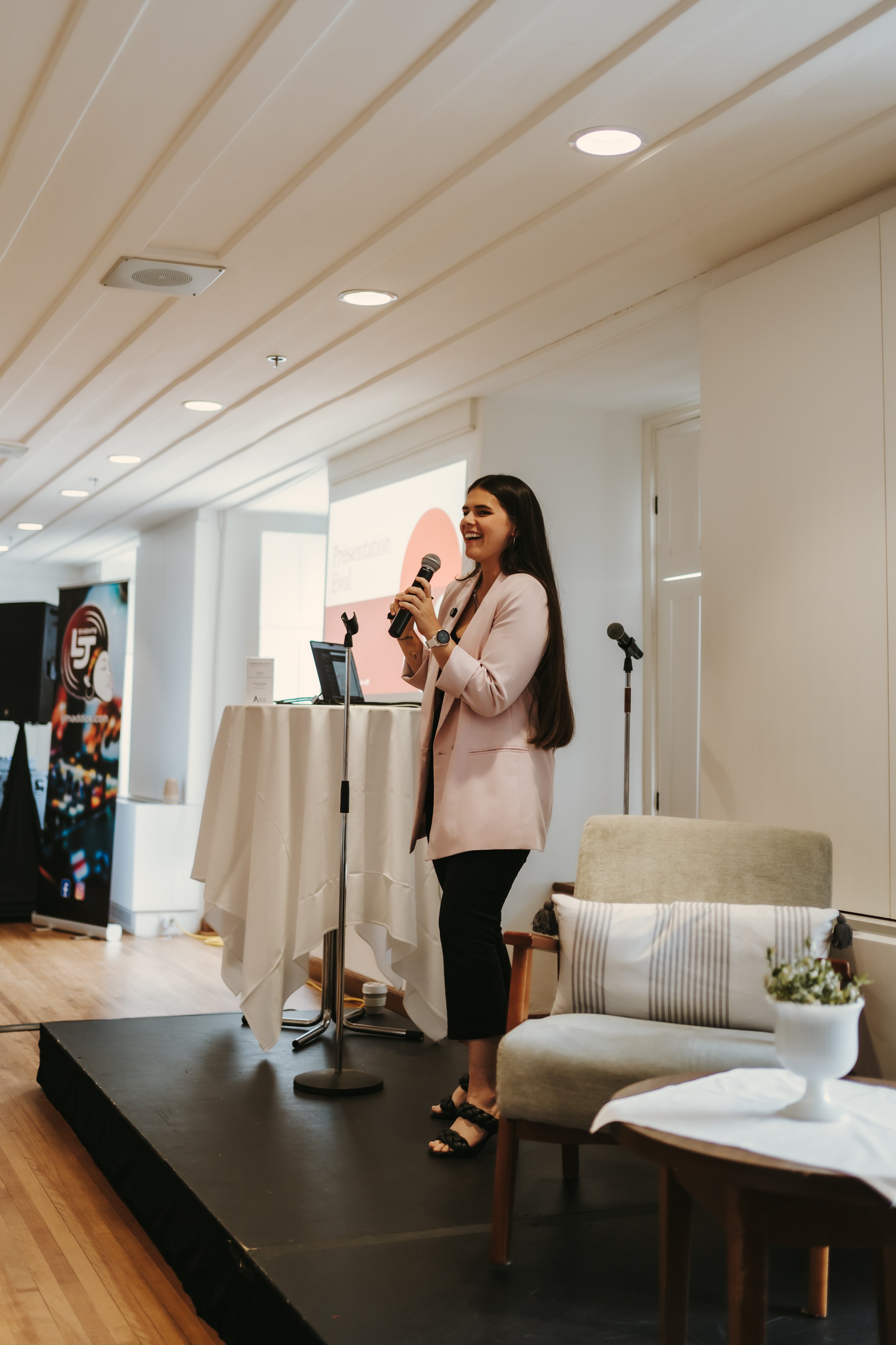 Woman speaking into a microphone on stage, standing beside a table with a laptop and projector screen in the background, in a conference room setting.