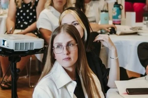 People attending a seated event, focusing on a woman with glasses. She appears to be listening attentively, with a projector and tables in the background.