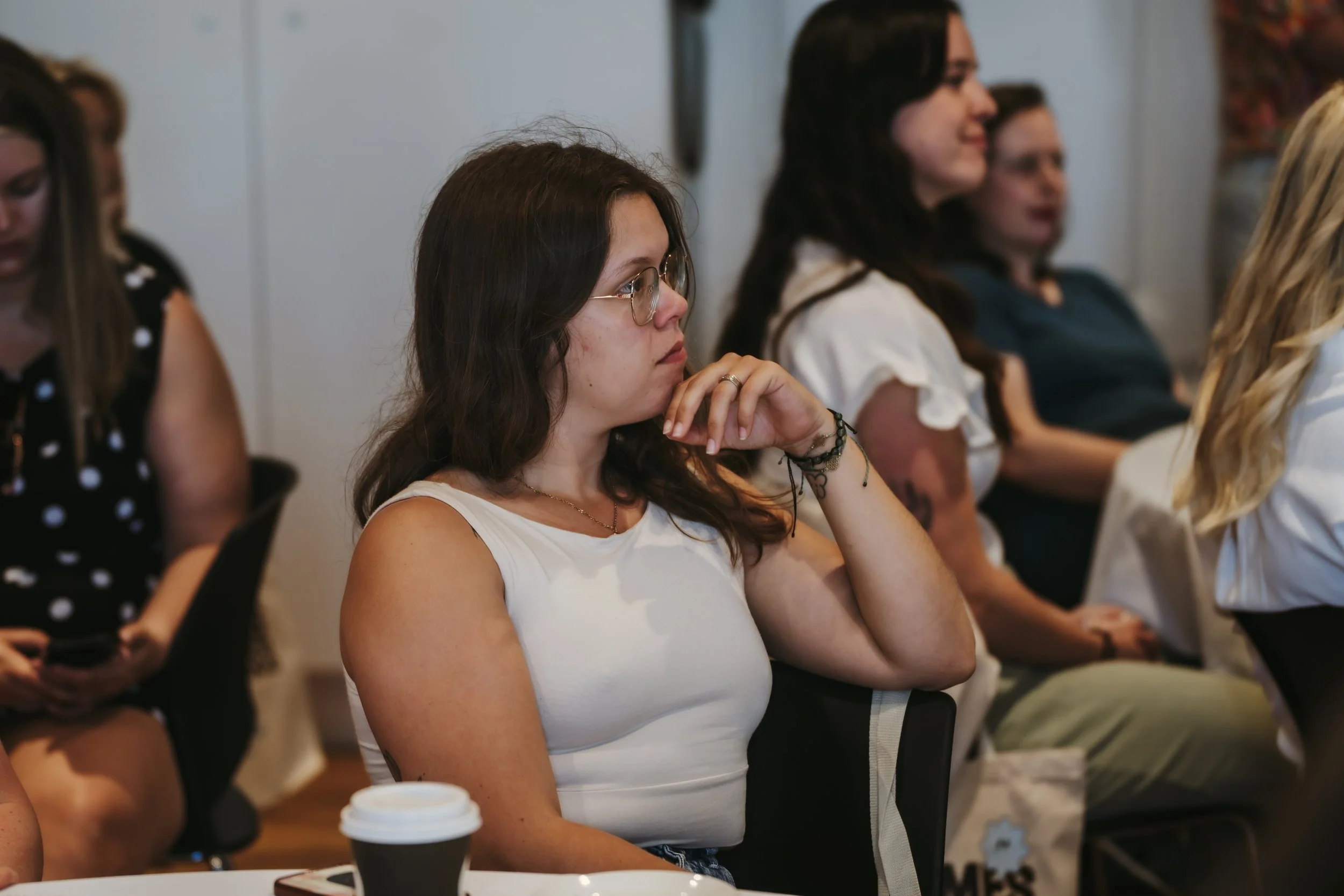 Groupe de personnes assises à l'intérieur, écoutant attentivement, avec un focus sur une femme en haut blanc et lunettes au premier plan.