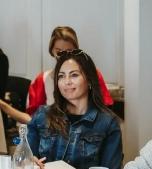 A woman with sunglasses on her head, wearing a denim jacket, sitting in a meeting room.