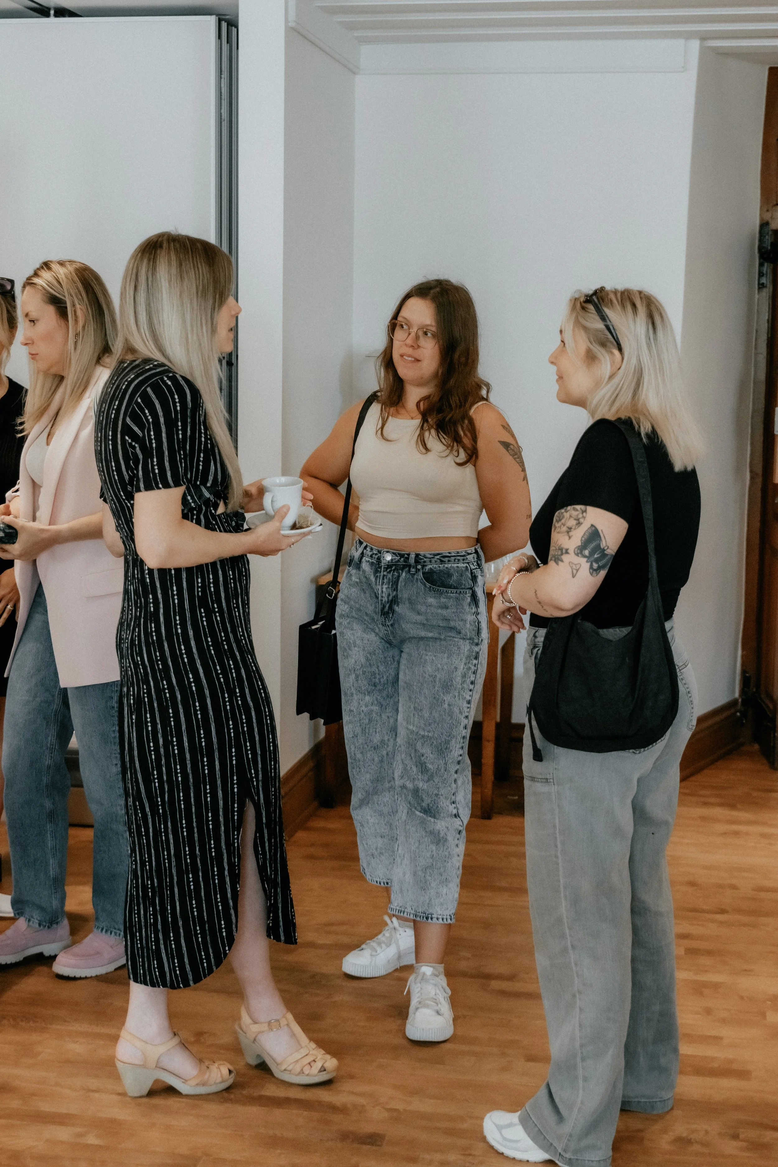 Group of women talking indoors, wearing casual clothing, one holding a cup and saucer.