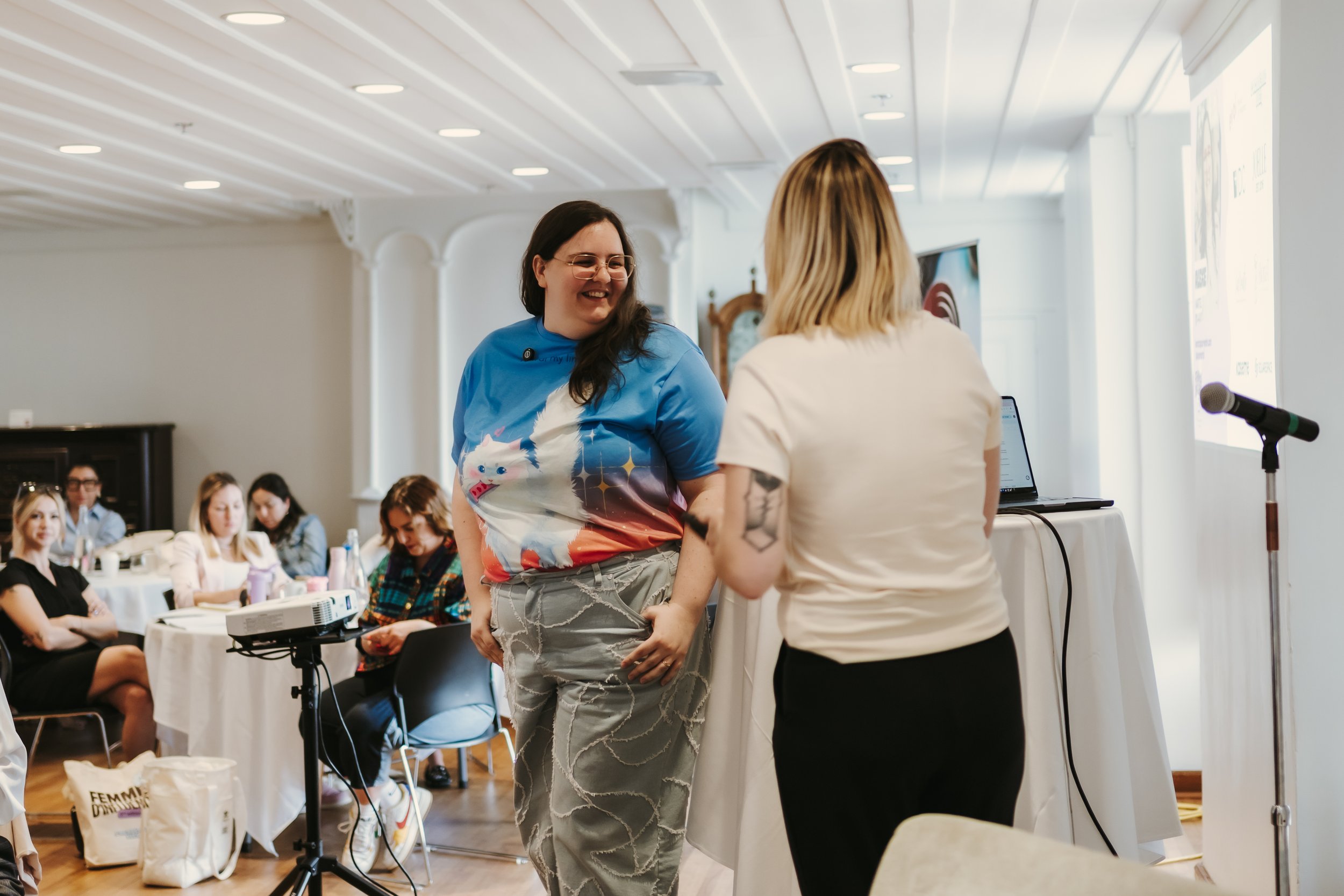 Two individuals conversing in a conference room with seated attendees. One person is wearing a blue shirt with a cat design, and the other has a tattoo. A laptop and microphone are visible on a table.