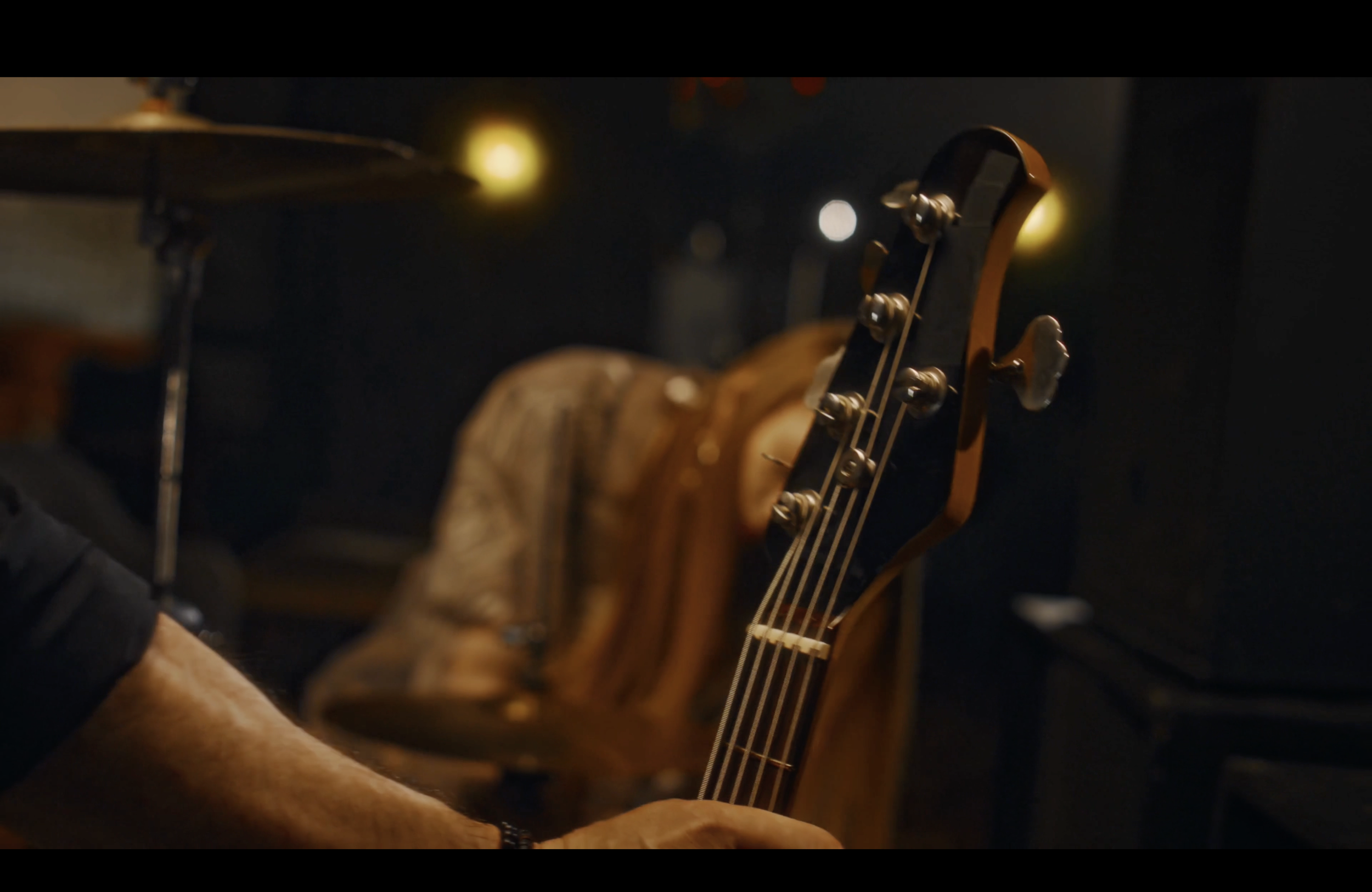 Close-up of a person's hand playing a black electric bass guitar in a dimly lit room with musical instruments in the background.