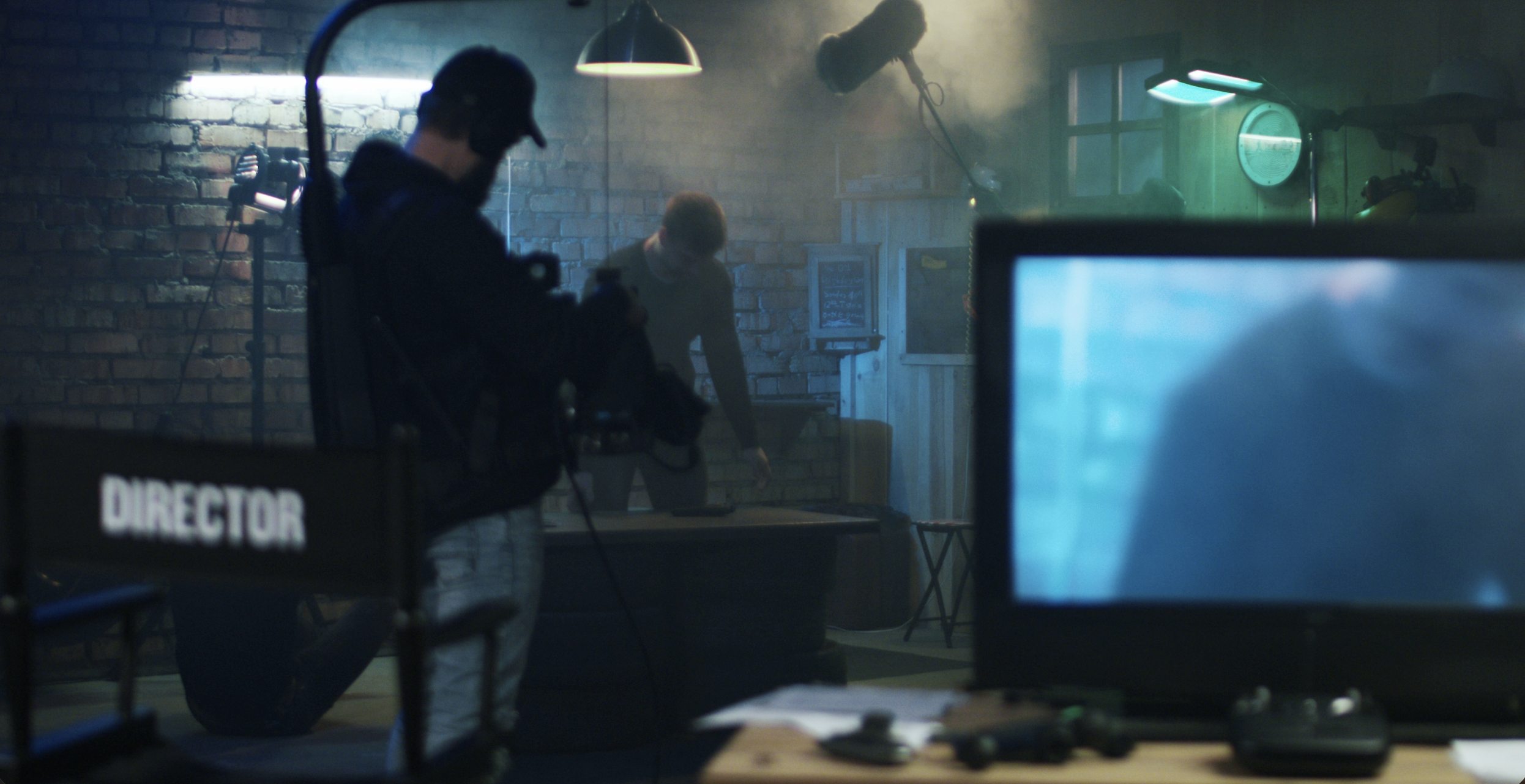 Clapperboard and director's chair on a film set with crew members and equipment, including a monitor and lighting, in a room with brick walls and a large window.