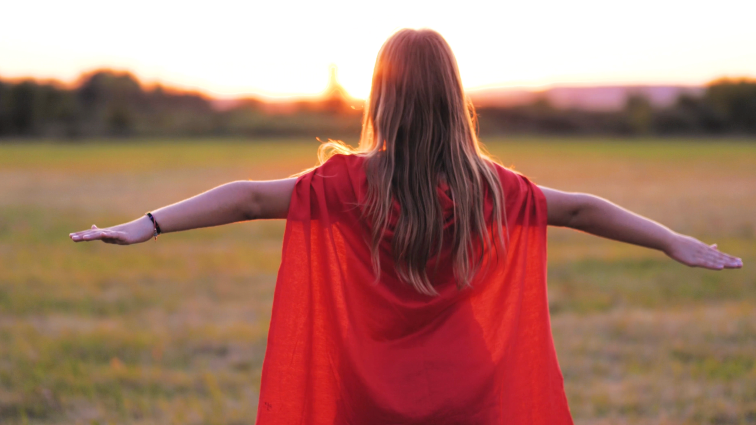 A woman with long red hair wearing a red cape stands in an open field at sunset with arms outstretched.