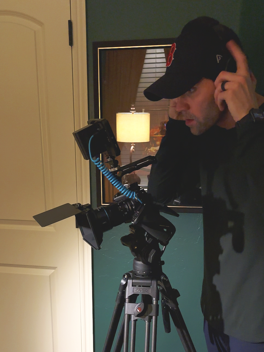 A man wearing a black cap and jacket using a professional video camera on a tripod inside a room, with a mirror reflecting a lamp and window behind him.