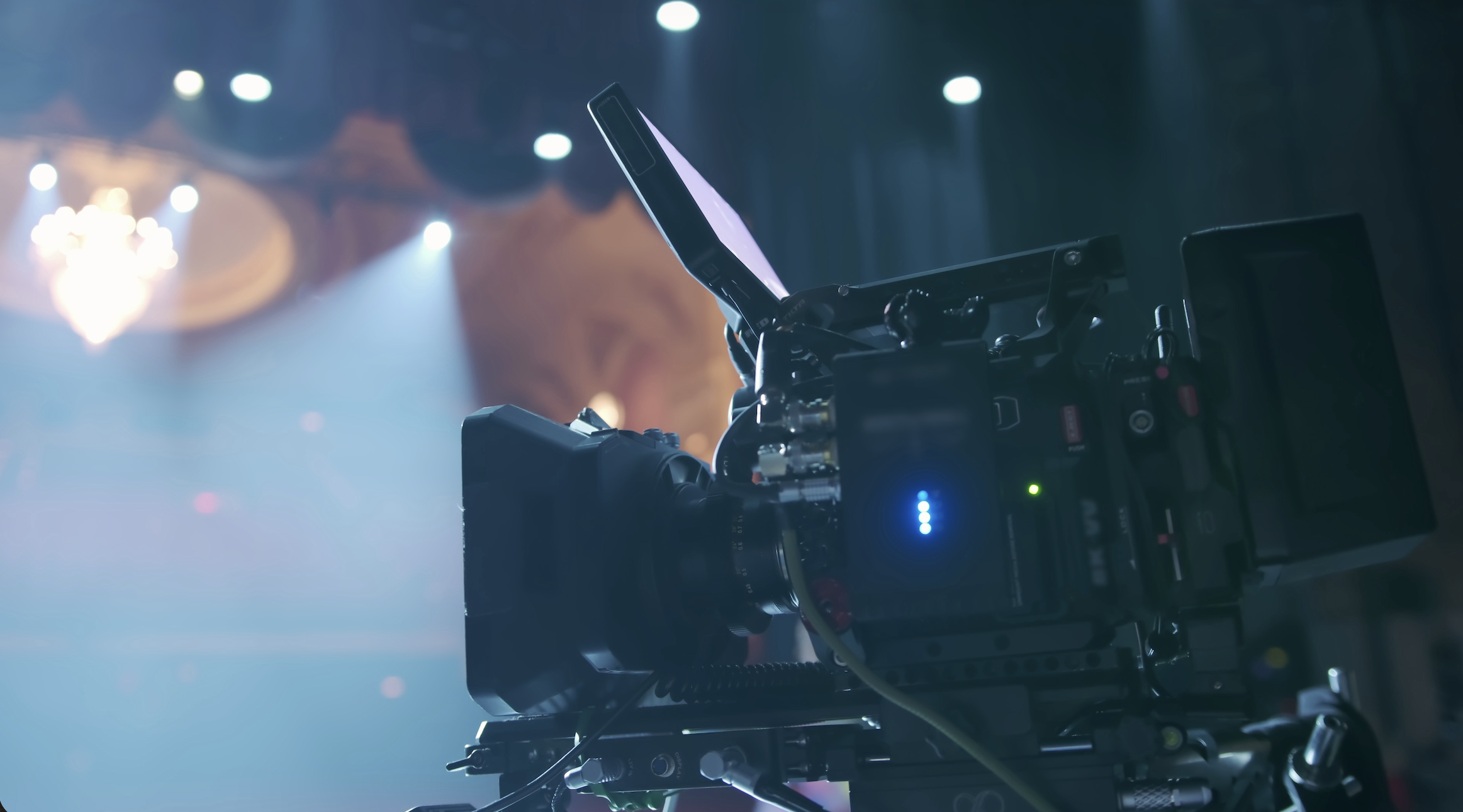 A professional video camera on a tripod in a dimly lit theater or stage setting.