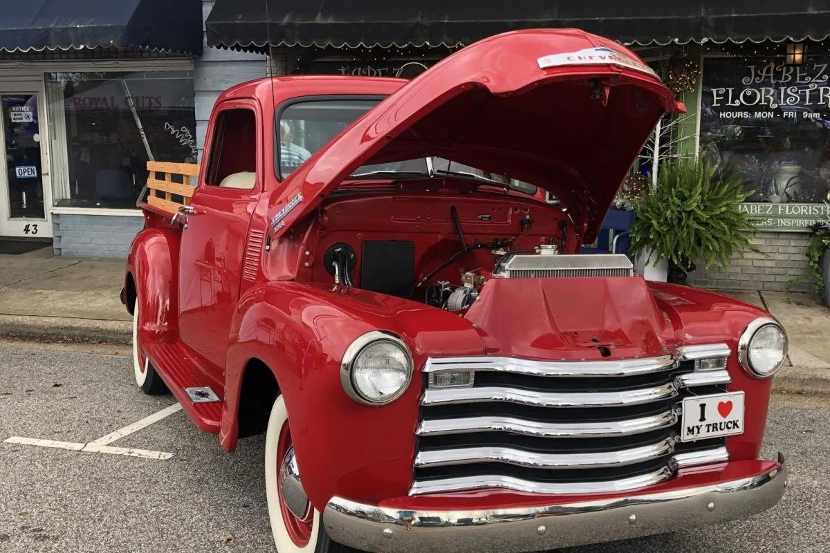 Vintage red Chevrolet pickup truck with hood open, parked in front of storefronts, showing engine and "I Love My Truck" license plate.