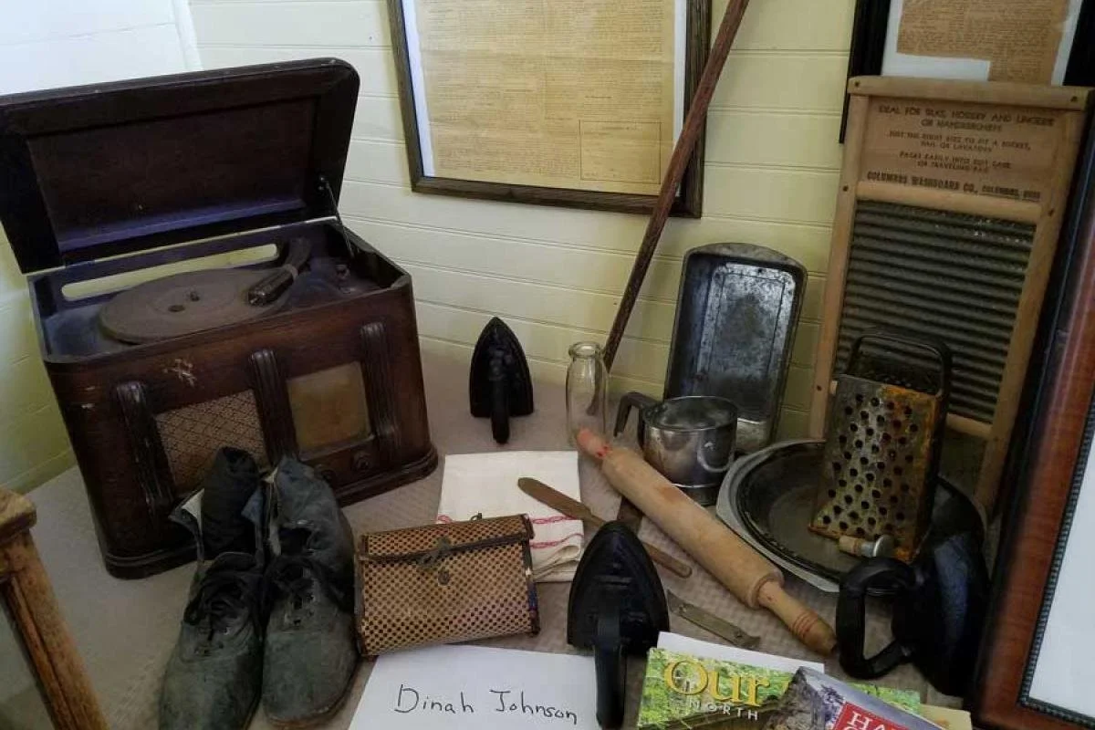 Vintage household items including a gramophone, old shoes, flat irons, a grater, a washboard, rolling pin, and other antique kitchen tools displayed on a table.