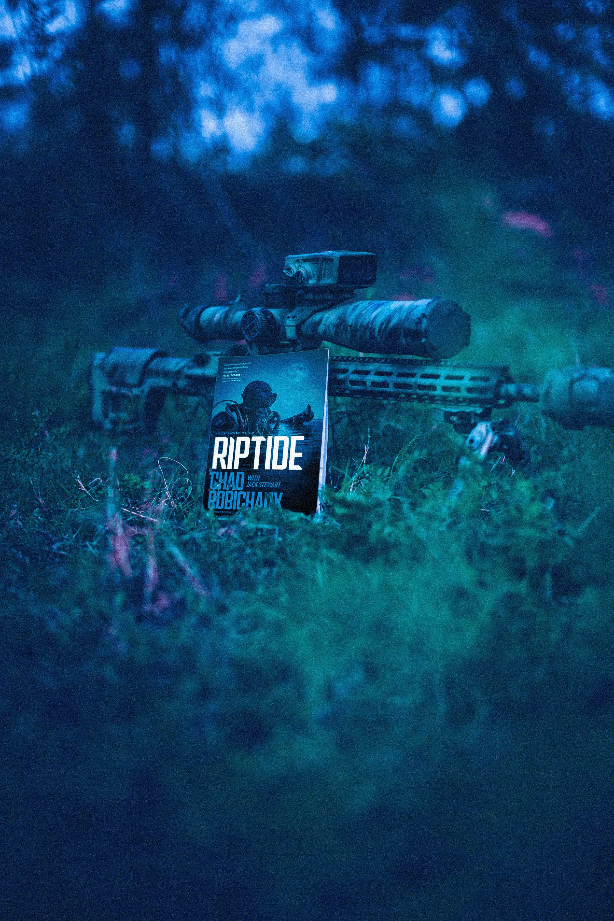 A book titled "RIPTIDE" standing against a camouflaged sniper rifle on grass.