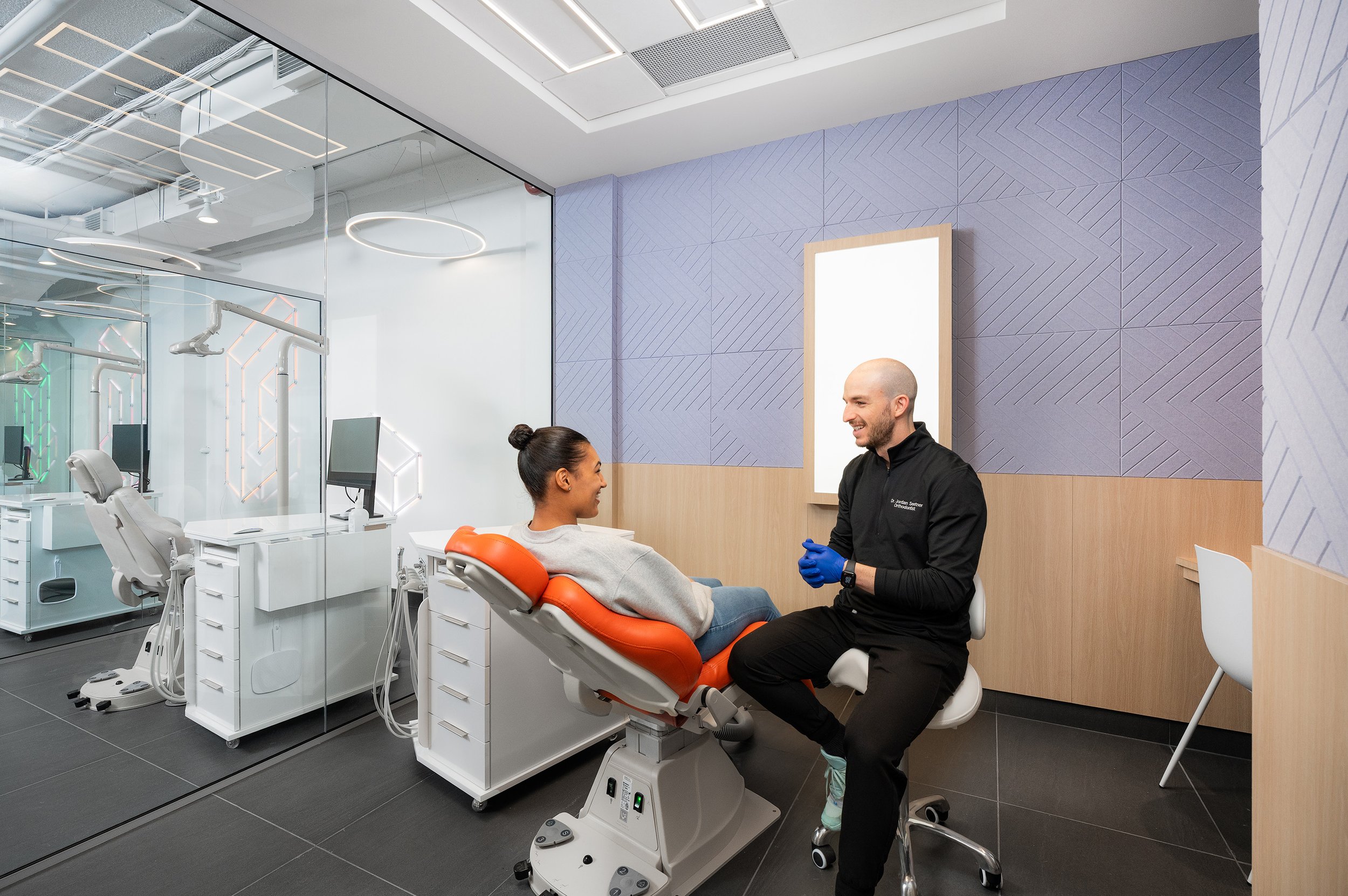 Dental office with a modern design, featuring a dental chair, glass partitions, digital equipment, and a dentist in conversation with a patient.