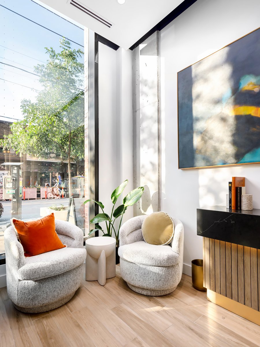 Modern interior space with two cushioned armchairs, one with an orange pillow and the other with a yellow pillow. A small round table is placed between the chairs. A large window with a view of the street and a potted plant beside the chairs is visible. On the wall, there is an abstract painting and a black console table with books and decor items. The floor is wooden.