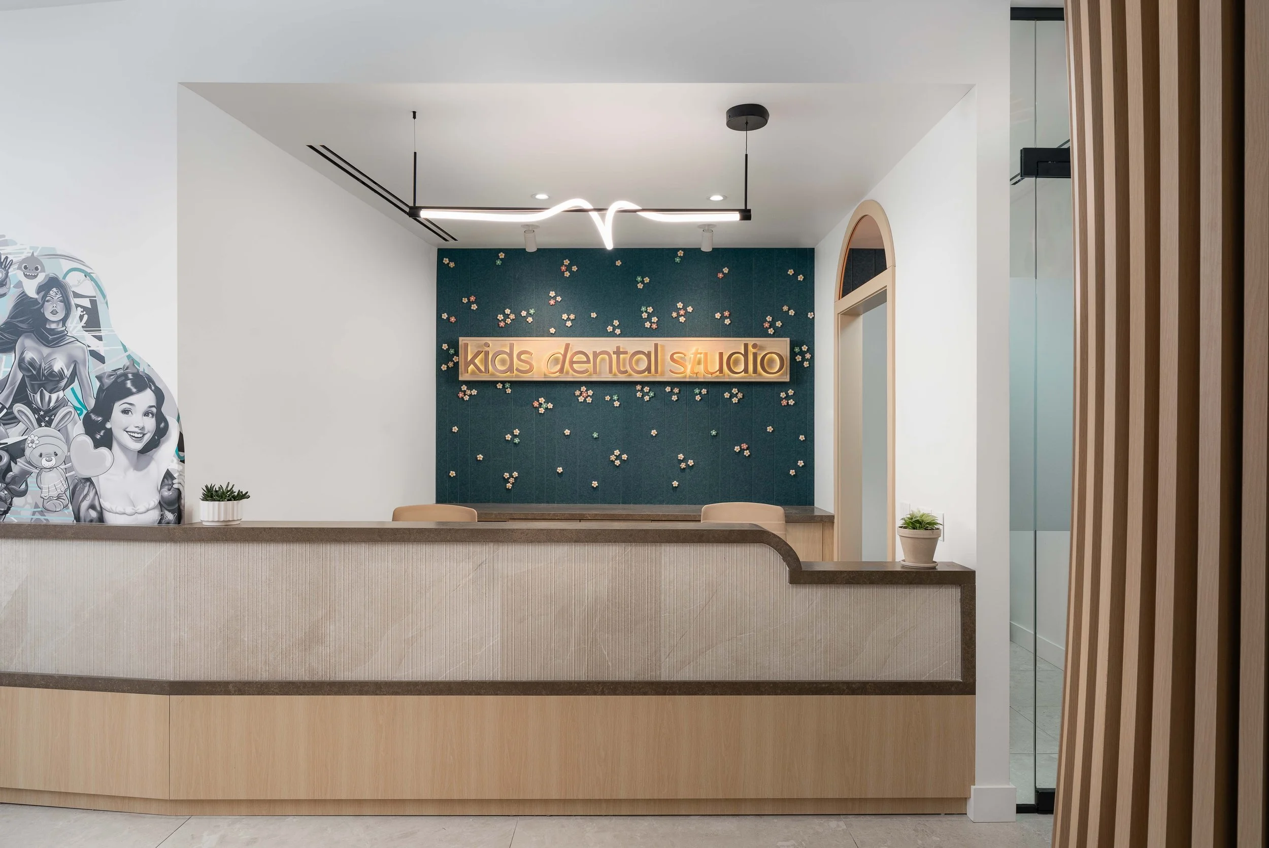 Reception area of a kids dental studio with a modern design, featuring a sign that says 'kids dental studio' on a decorative wall. Cartoon character images and potted plants are visible on the counter and wall.