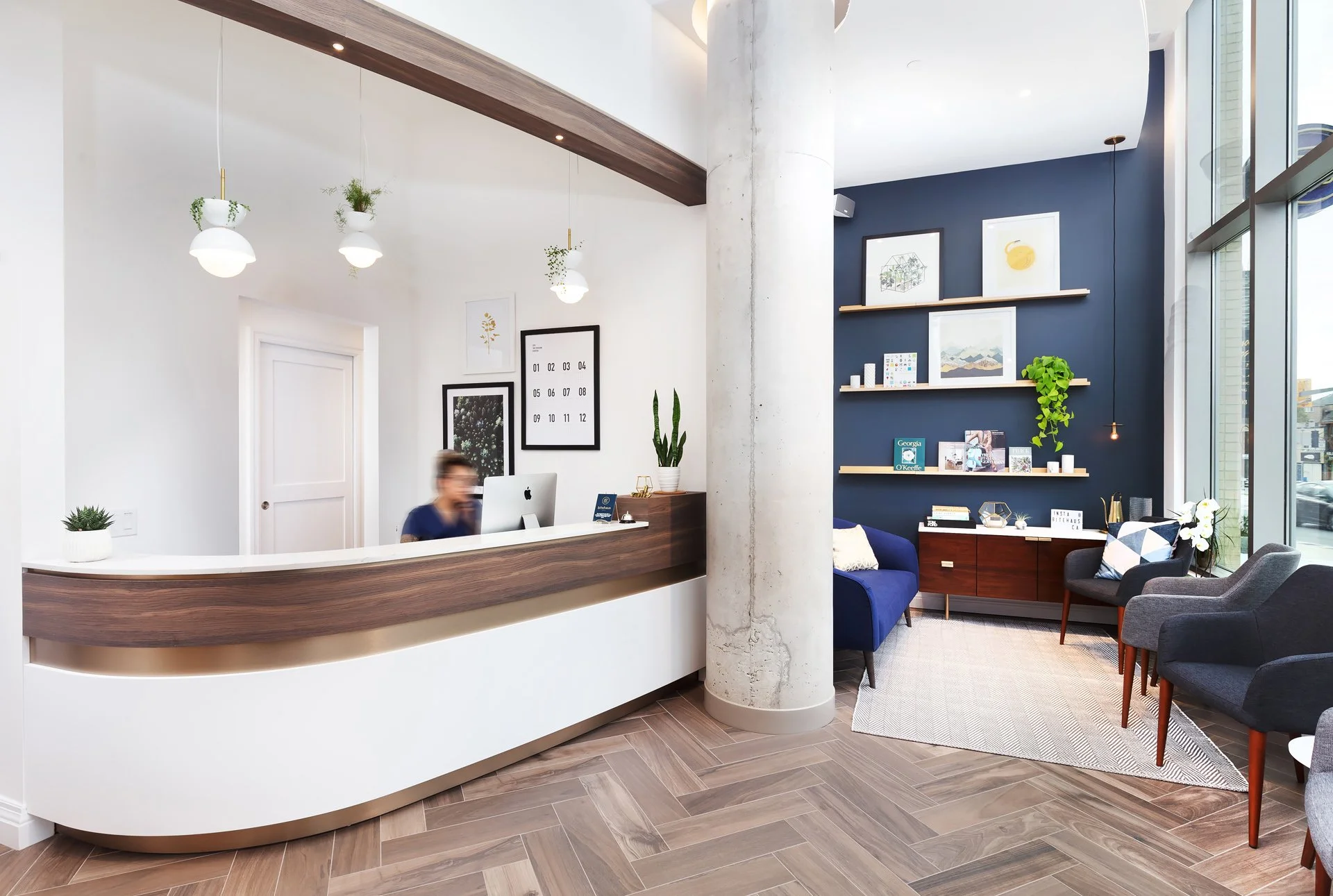 Modern reception area with a white and wood desk, a person working behind it, minimalist decor, navy blue wall with shelves, plants, and framed art, alongside a seating area with armchairs and a rug.