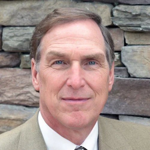 A man with long dark hair, sunglasses, and a beard wearing a light blue shirt leaning against a stone wall, looking at his smartphone.