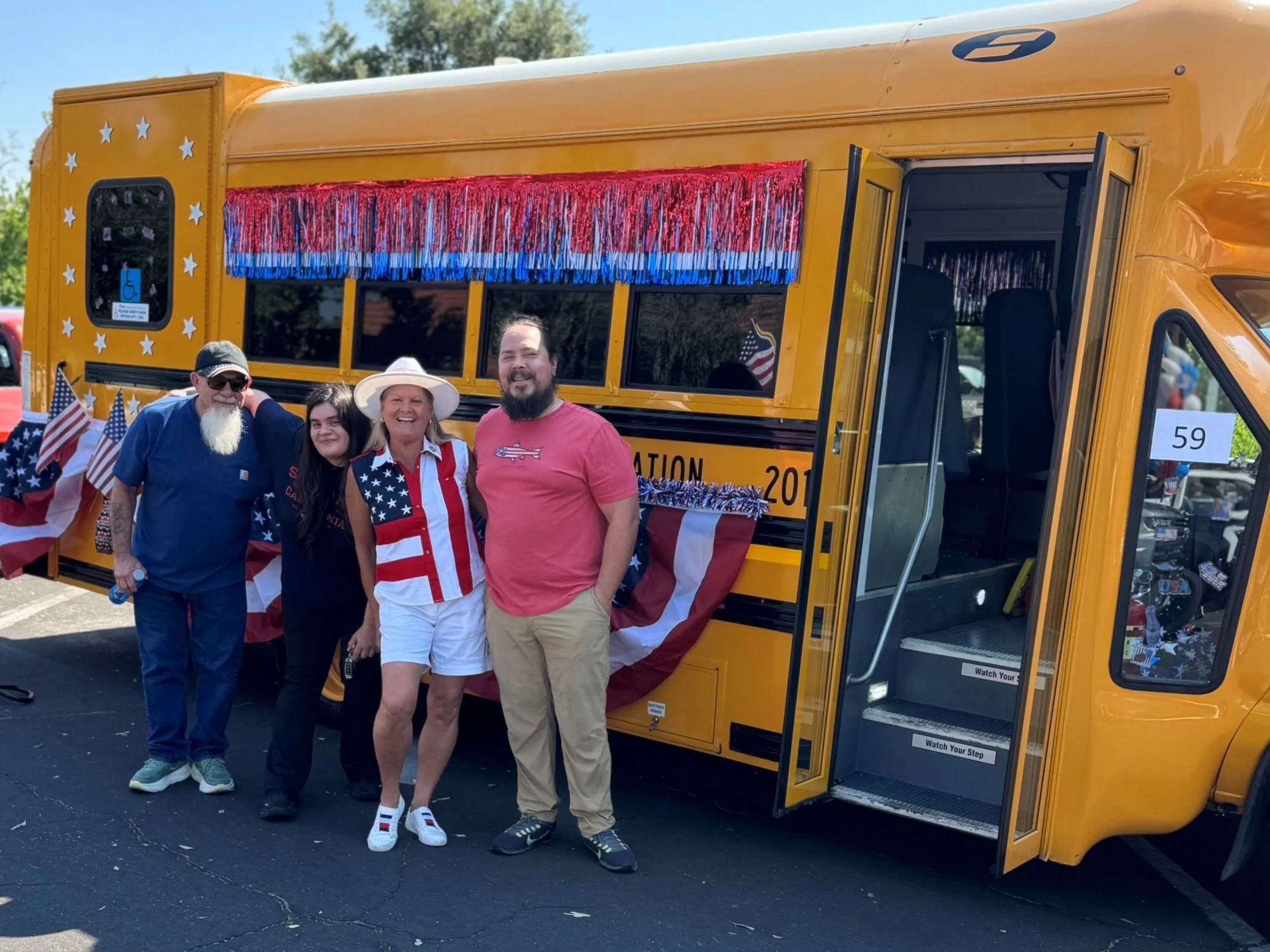 Four people standing in front of a decorated yellow bus with American flag decorations, outdoors on a sunny day.