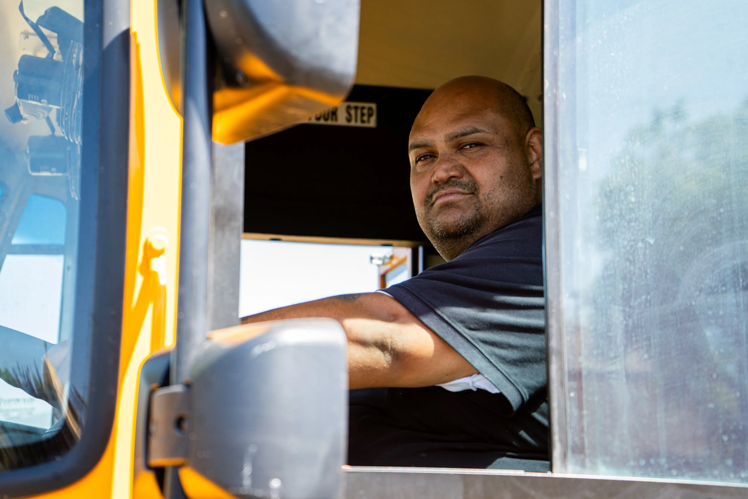 A man sitting in the driver's seat of a yellow truck, looking out the window with a serious expression.