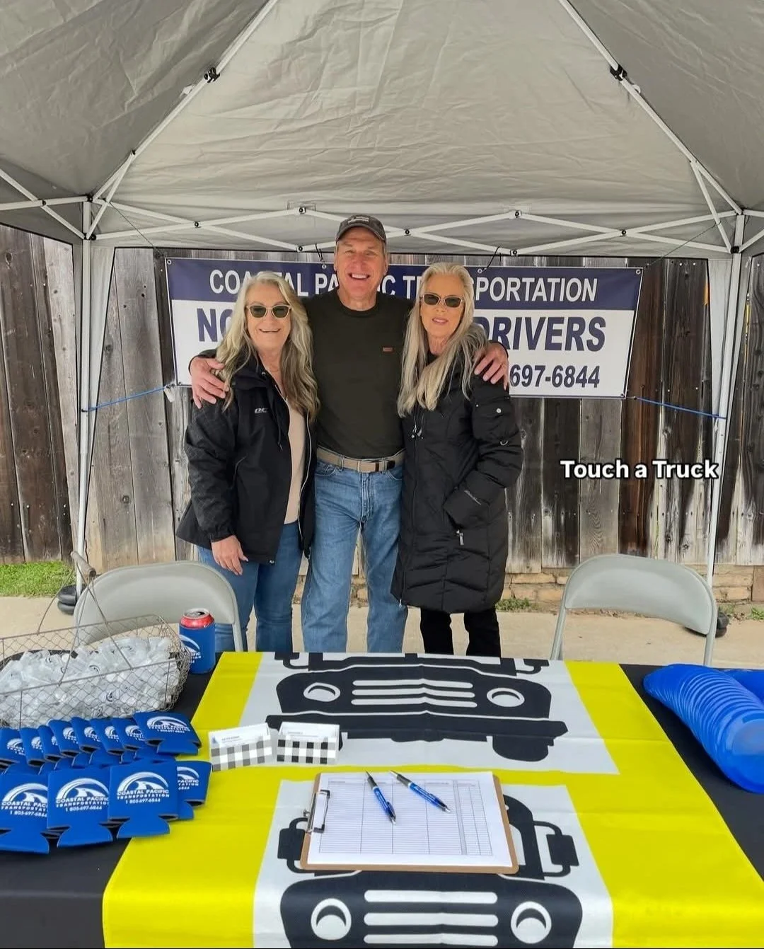 Three smiling people standing under a canopy at a community event, with a table in front of them that has a yellow tablecloth and clipboards, and a banner behind them that indicates the event is related to coastal Pacific transportation and no contac