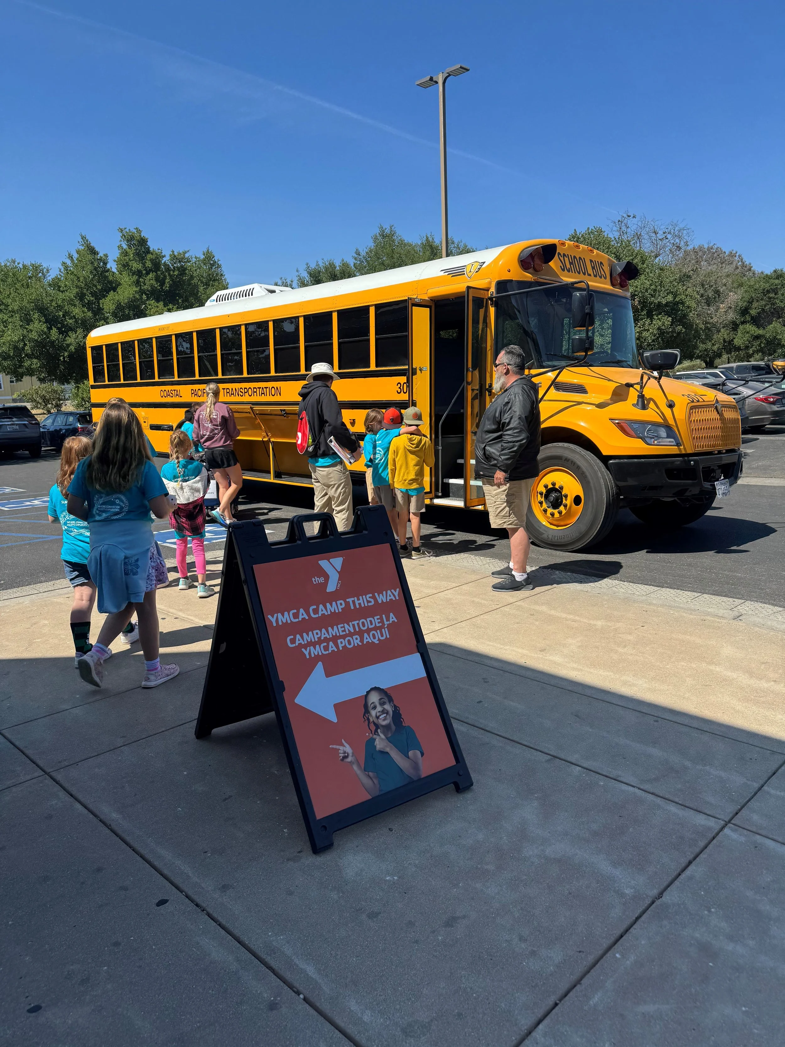Children and adults boarding a yellow school bus outside a parking lot on a sunny day, with a sign directing to YMCA camp