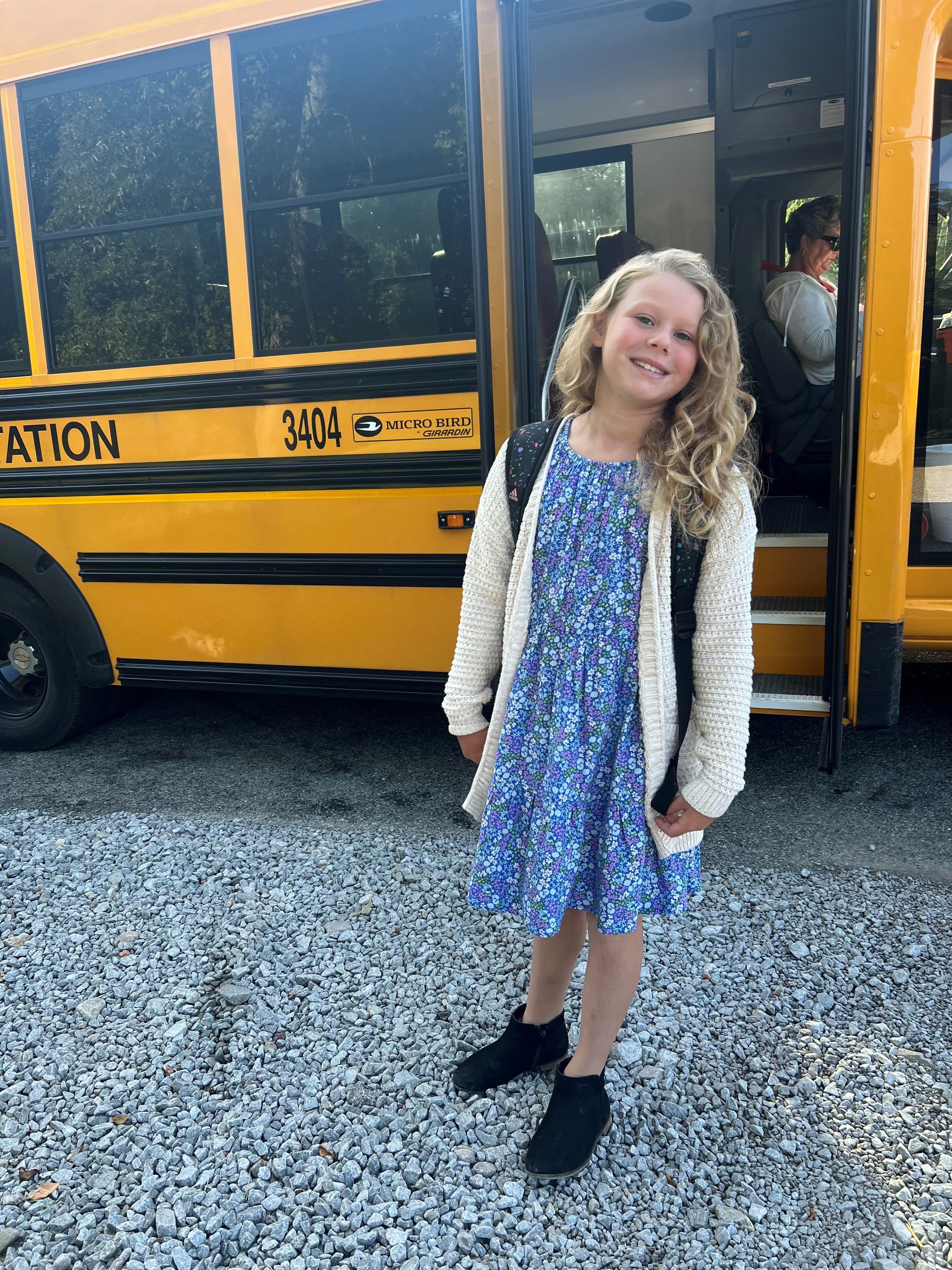 Young girl with curly blonde hair smiling and standing in front of a yellow school bus, wearing a floral dress, a white cardigan, and black boots, with a backpack on her shoulders.