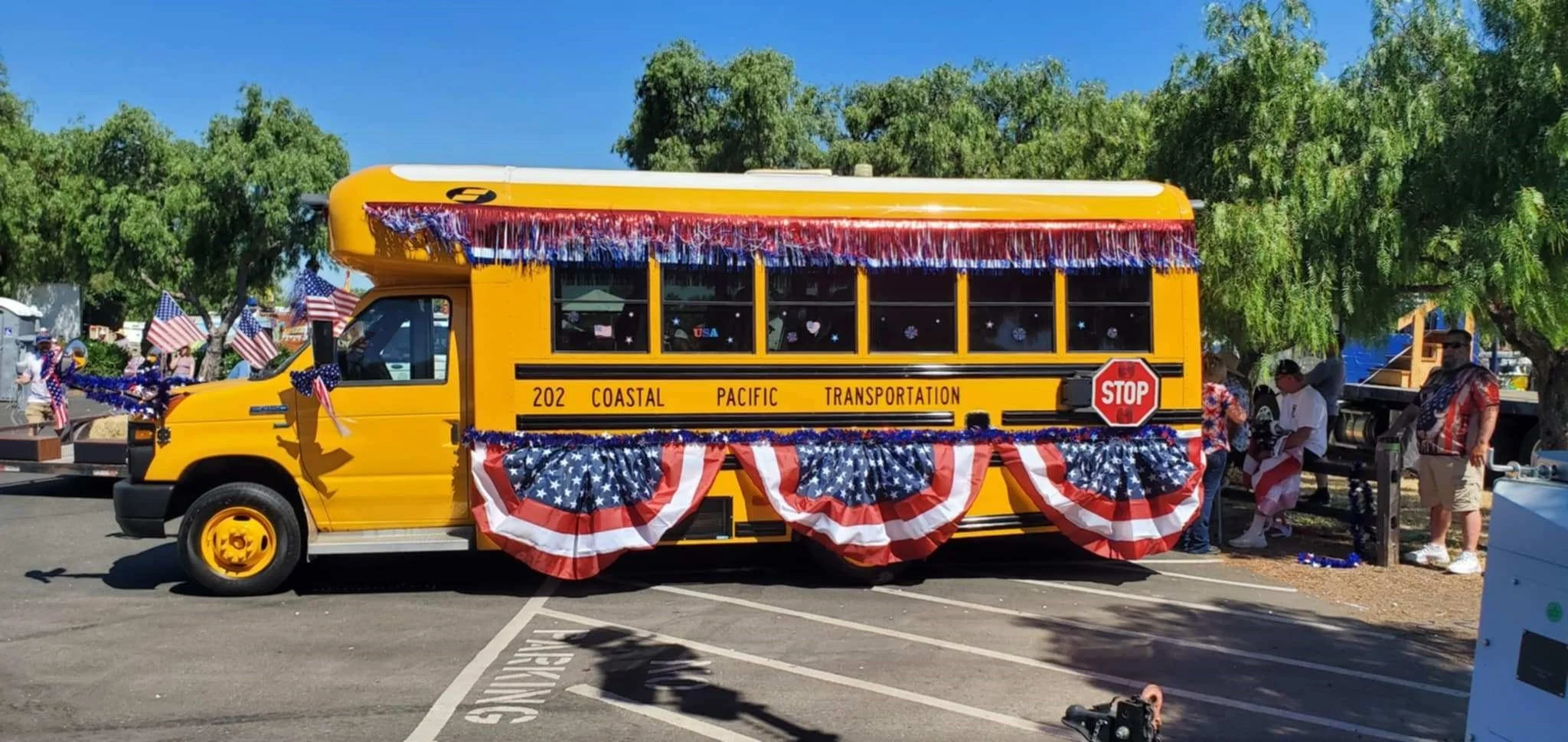 A decorated yellow school bus with patriotic decorations, including American flags and red, white, and blue bunting, parked in a lot with people around it, during a parade or celebration.
