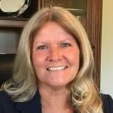 Portrait of a smiling woman with shoulder-length blonde hair, wearing a dark top, standing in front of a wooden cabinet.