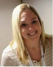 A young woman with blonde hair wearing a sleeveless blue denim or chambray shirt, smiling and sitting on a brown leather bench or sofa.