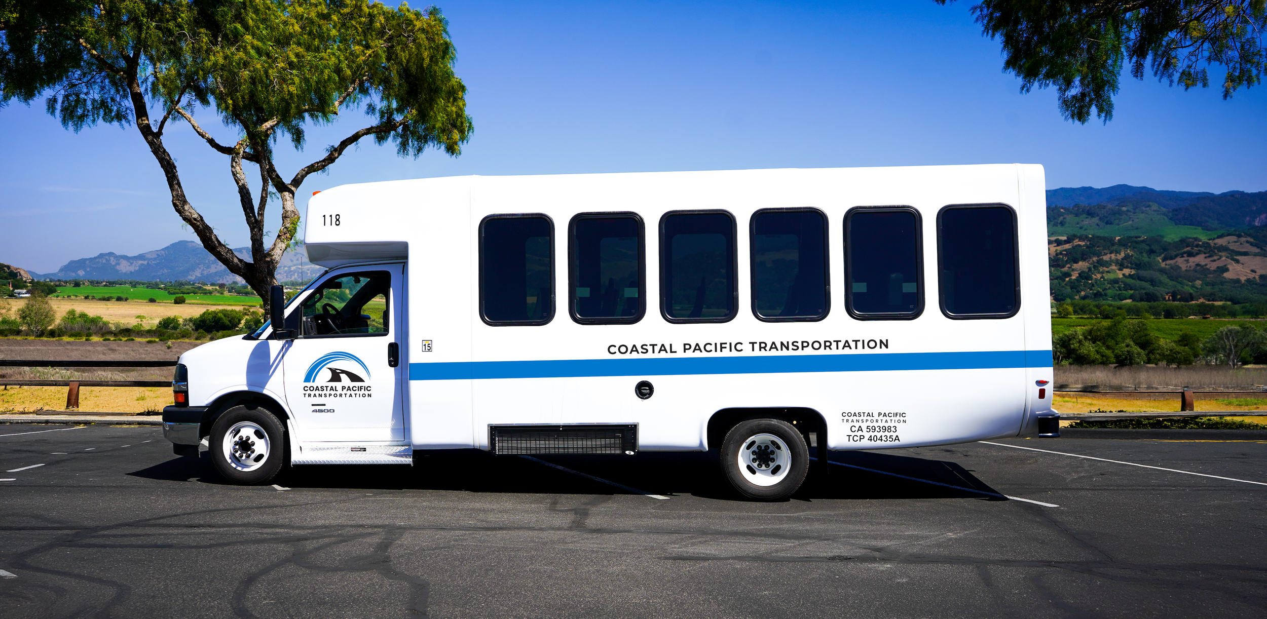 A white shuttle bus with blue accents and the words 'Coastal Pacific Transportation' parked in an outdoor lot with mountains, green fields, trees, and a clear blue sky in the background.