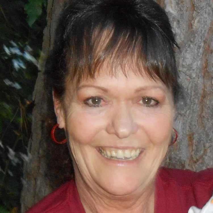 A woman with dark hair, wearing a black fuzzy sweater and white pants, sitting on a striped blanket on a couch, smiling and looking to her right.