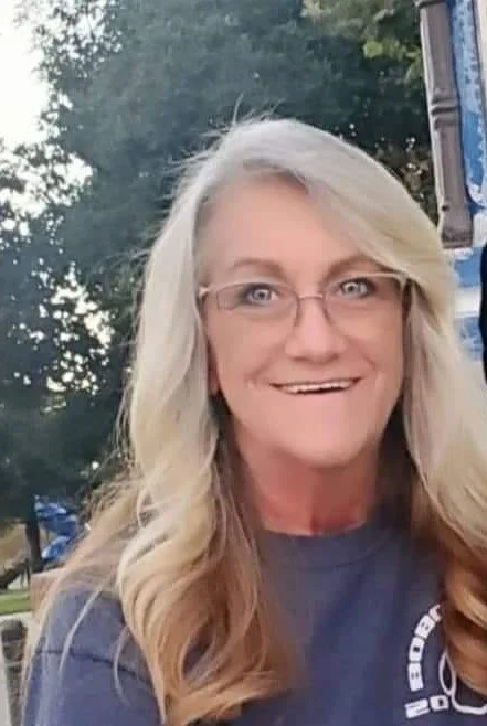 A woman with shoulder-length dark hair wearing a black sweater and white pants sitting on a striped couch, smiling and looking to her left.