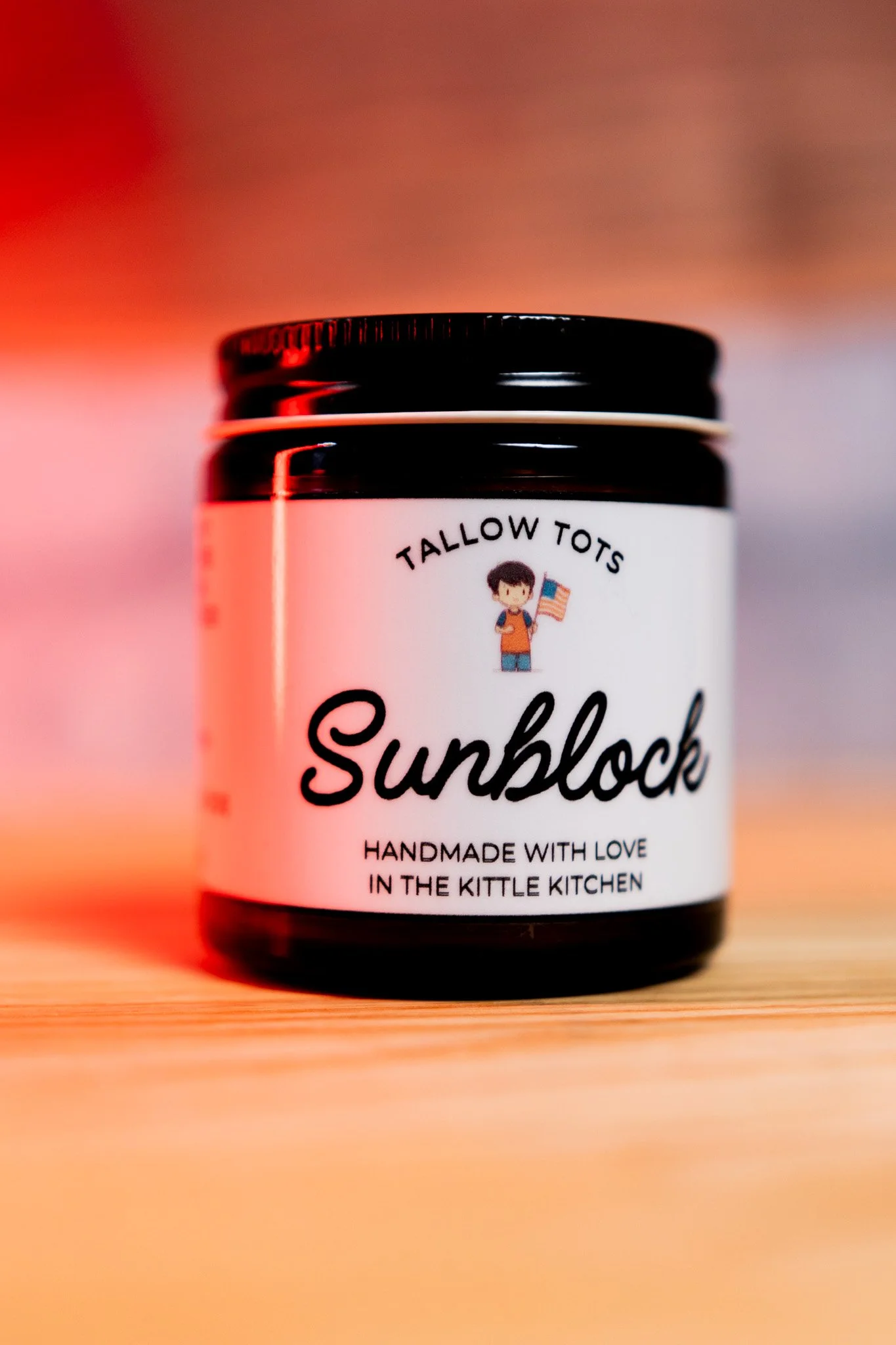 A black jar labeled "Sunblock" with a cartoon child holding an American flag, the brand name "Tallow Tots", and the phrase "Handmade with love in the Kittle Kitchen" on a wooden surface with a blurred pinkish background.