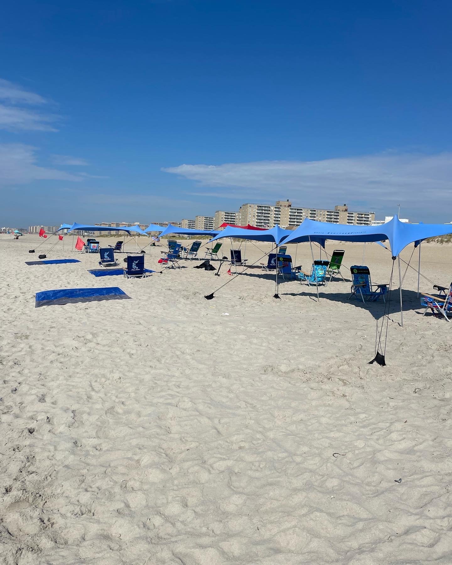 Beach with blue and red umbrellas, chairs, and towels on sandy shore, with a cityscape of tall buildings in the background under a blue sky with some clouds.