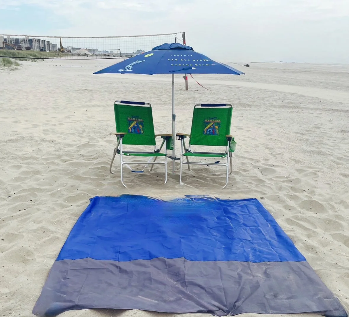 Beach scene with two green chairs and a large blue umbrella, a blue and gray blanket on the sand, and buildings in the distance.