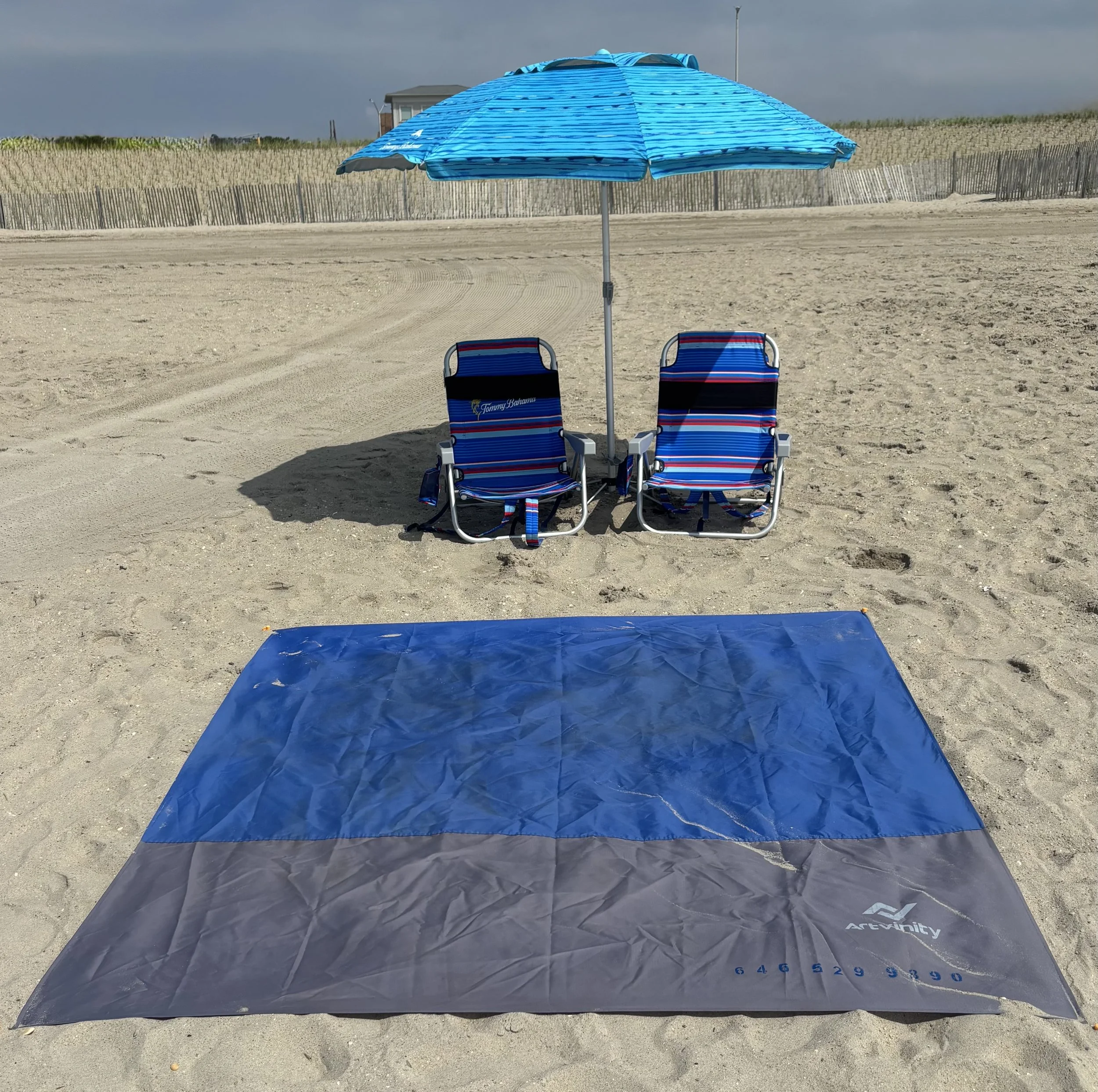 Beach scene with two striped blue and red chairs under a blue umbrella, and a blue and gray beach mat on the sand.