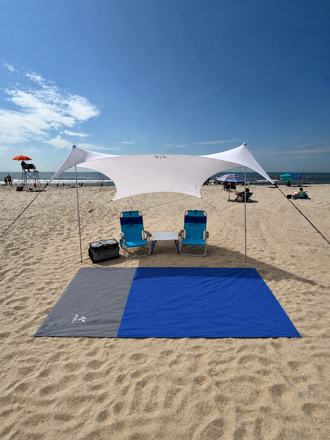 Beach setup with a blue shade tent, two blue folding chairs, a cooler, and a blue mat on sandy beach with the ocean in the background.