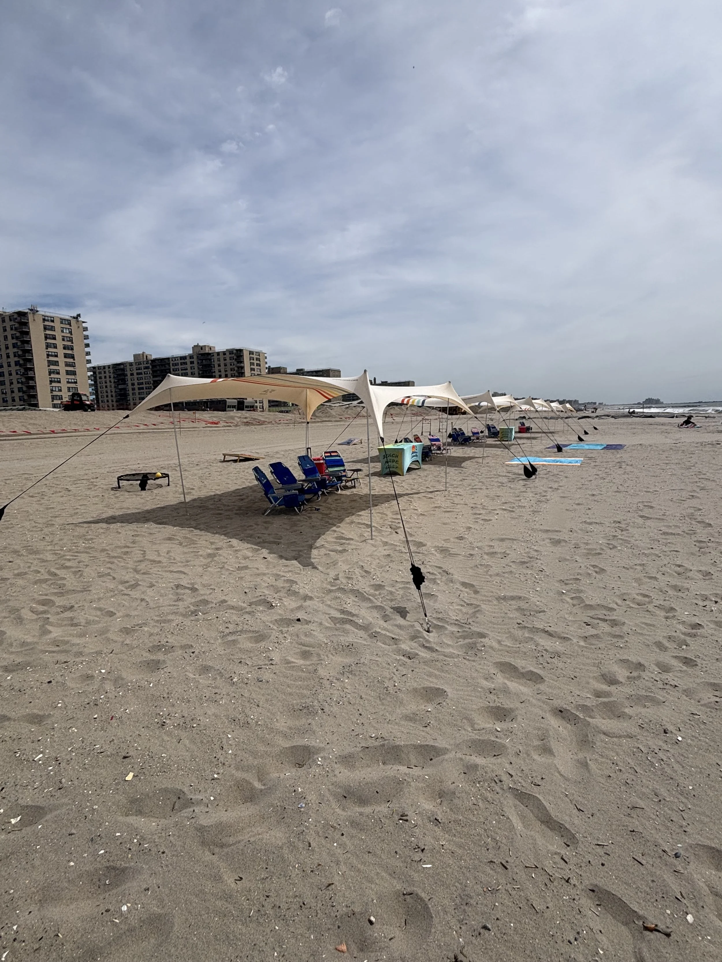 Empty beach with blue and red tents, chairs, and umbrellas set up on sandy shore, with a clear blue sky and buildings in the background.
