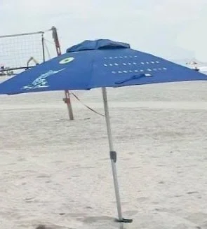 A blue beach umbrella on the sand with the ocean and a volleyball net in the background.