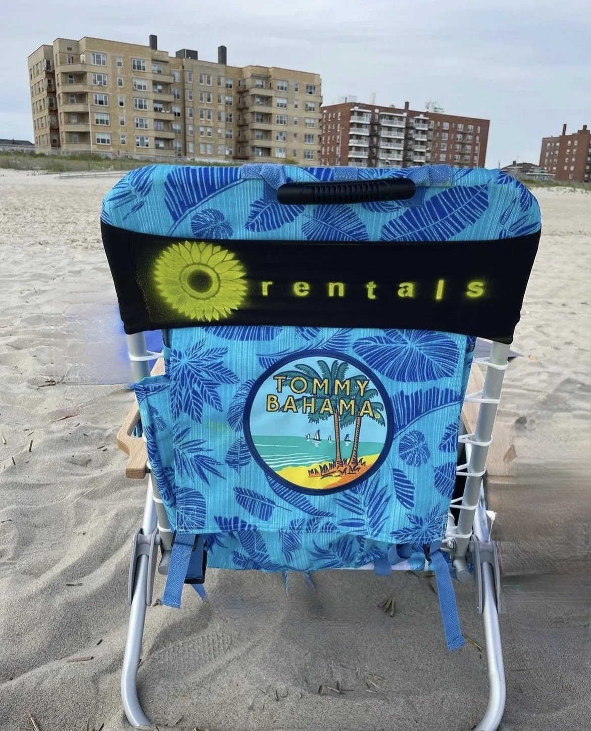 Beach chair with Tommy Bahama logo and a tropical design, situated on sandy beach with multi-story buildings in the background.