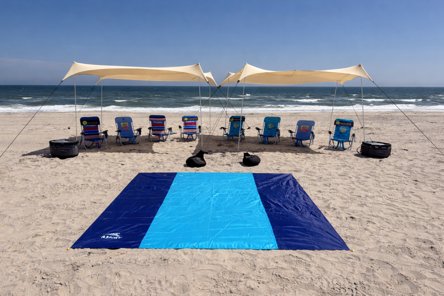 A beach setup with four chairs under two canopies, a blue and black blanket on the sand, and various small tables and belongings, overlooking the ocean with gentle waves and a clear blue sky.