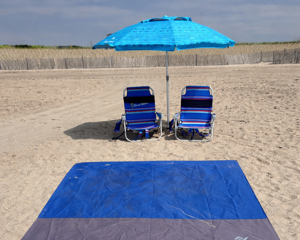 Beach scene with two green chairs under a blue umbrella and a blue and gray beach blanket on the sand, with buildings and cloudy sky in the background.