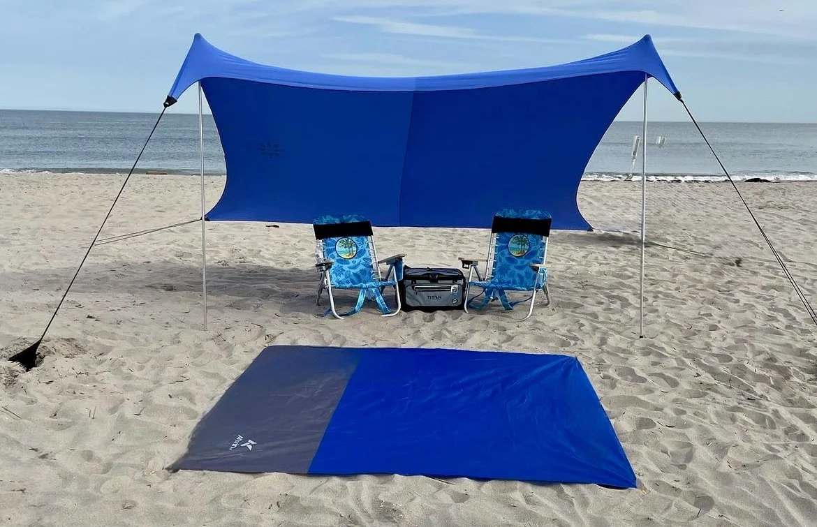 Beach setup with a blue shade tent, two blue folding chairs, a cooler, and a blue mat on sandy beach with the ocean in the background.