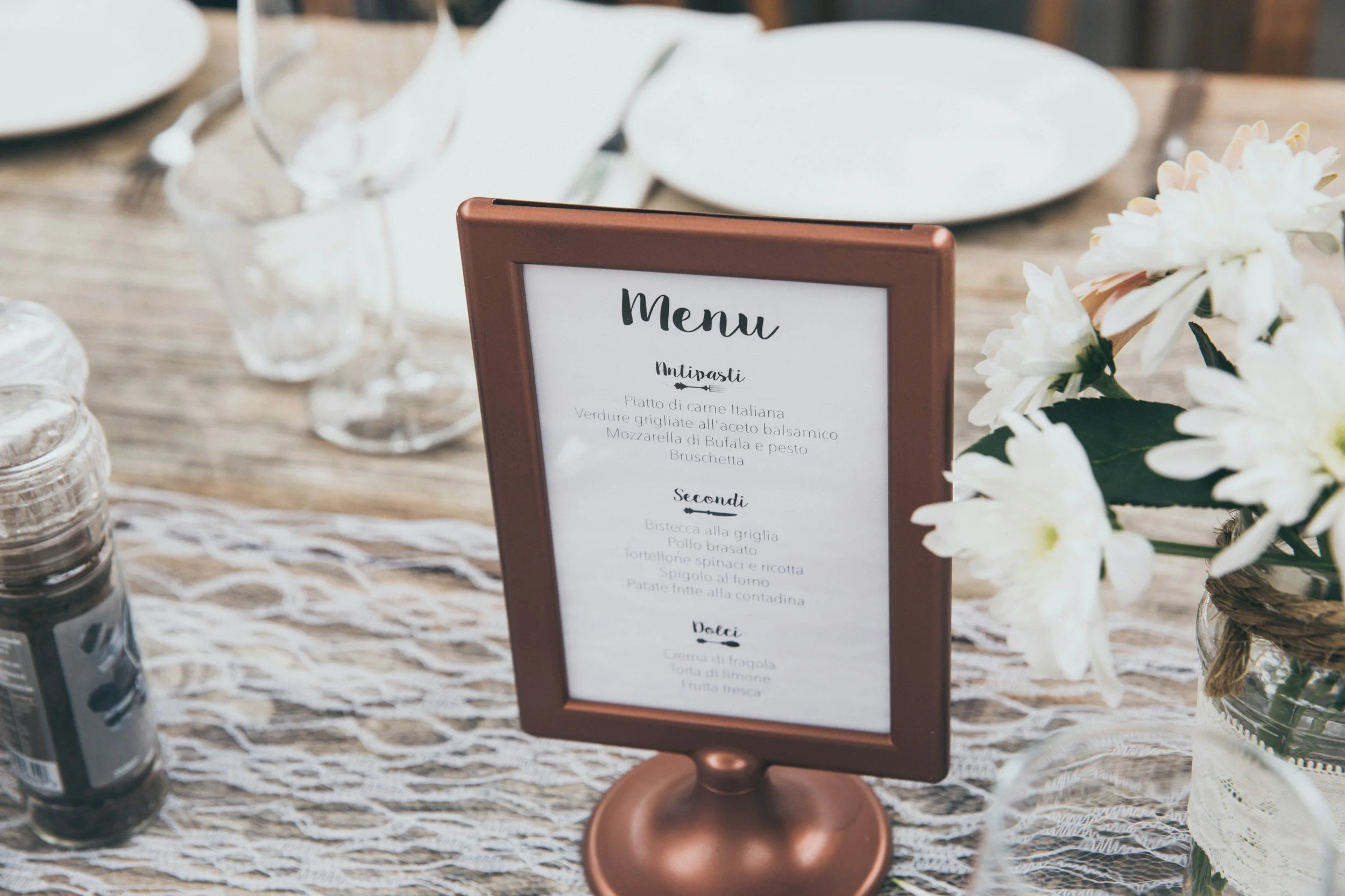 Menu on a table with white plates, glasses, and flowers in a glass jar, featuring Italian dishes listed on the menu.