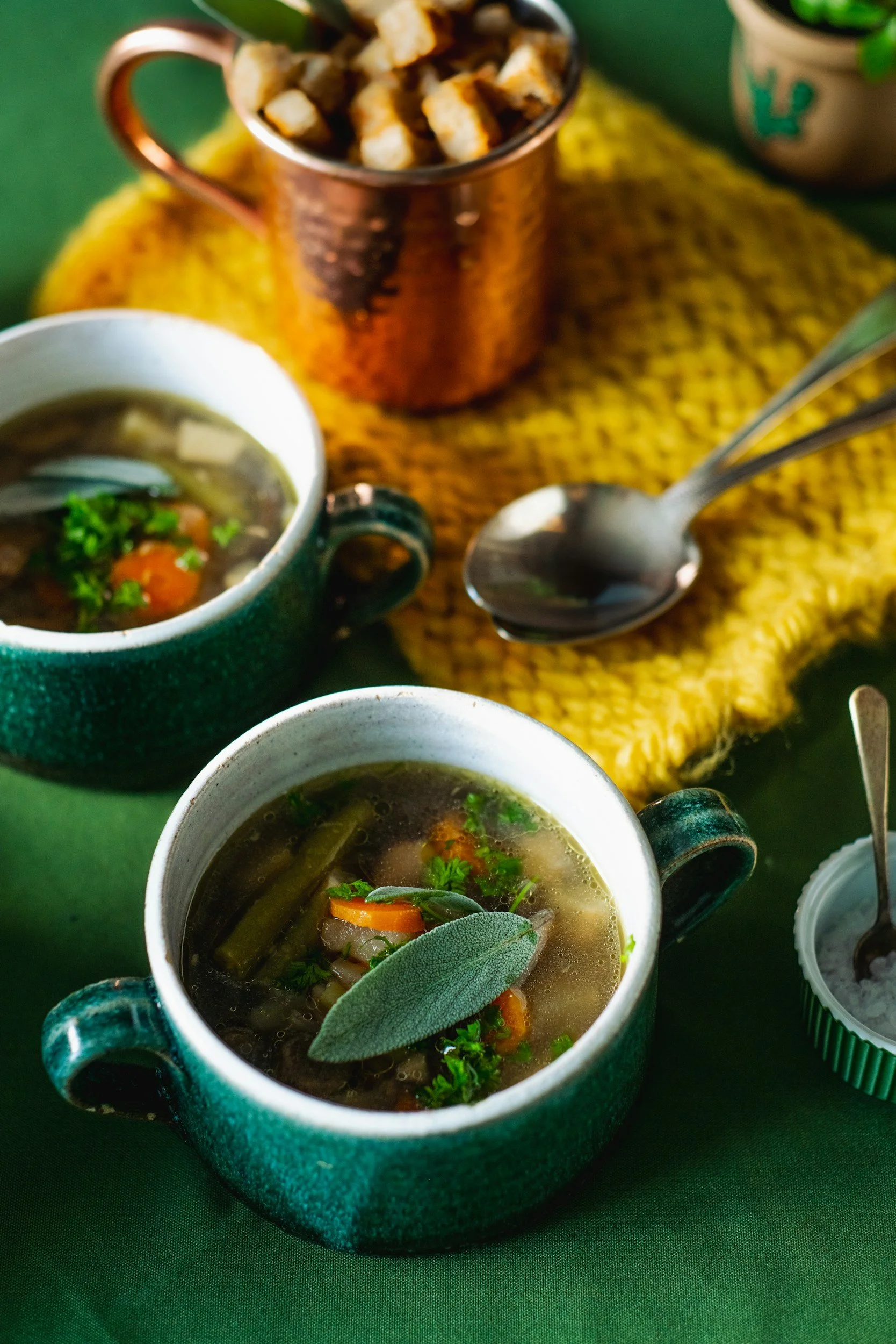 Two green ceramic cups of vegetable soup with herbs, a yellow cloth napkin, a spoon, a copper cup with croutons, and a small jar of salt on a green table.
