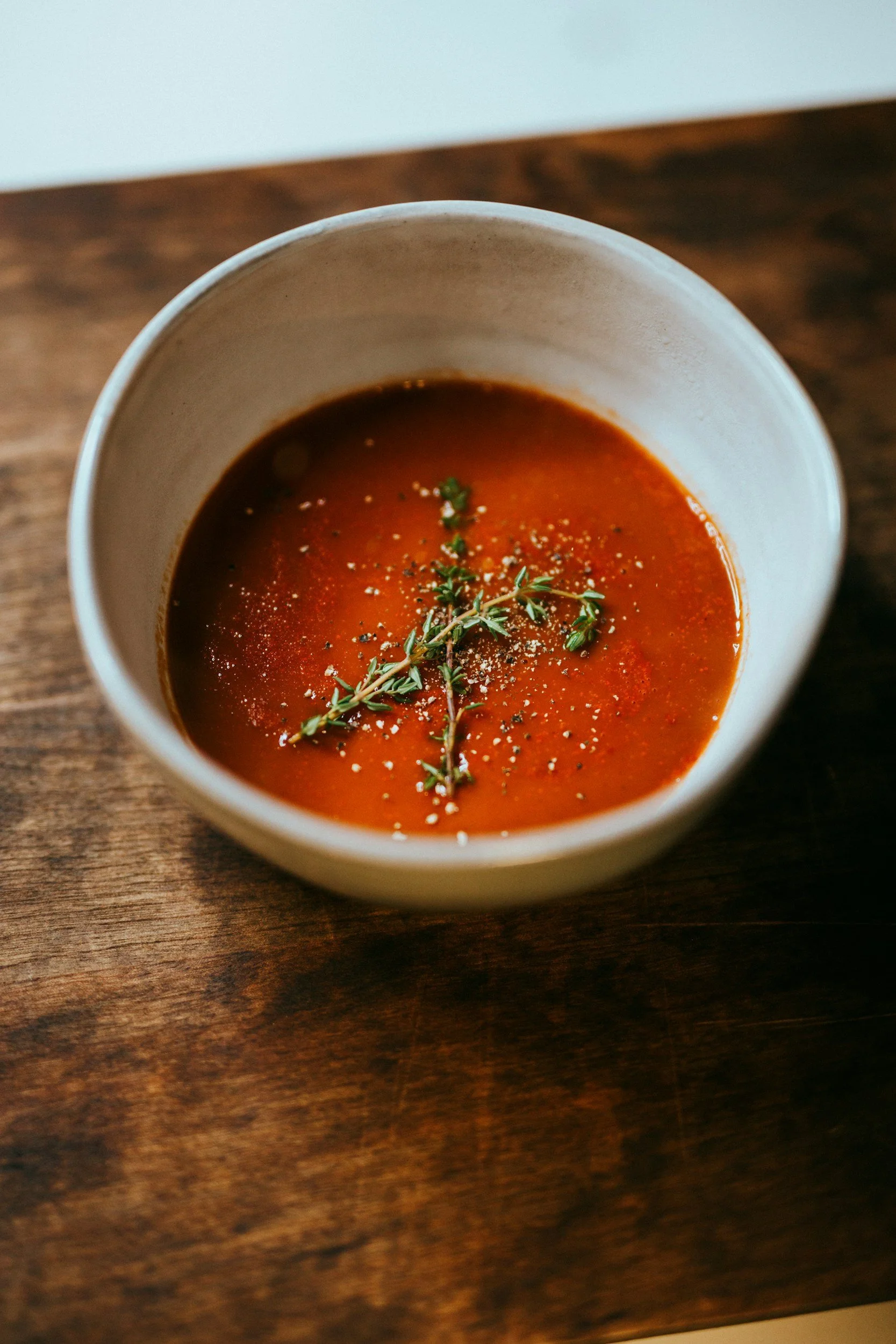 A bowl of tomato soup garnished with a sprig of thyme, black pepper, and salt on a wooden surface.