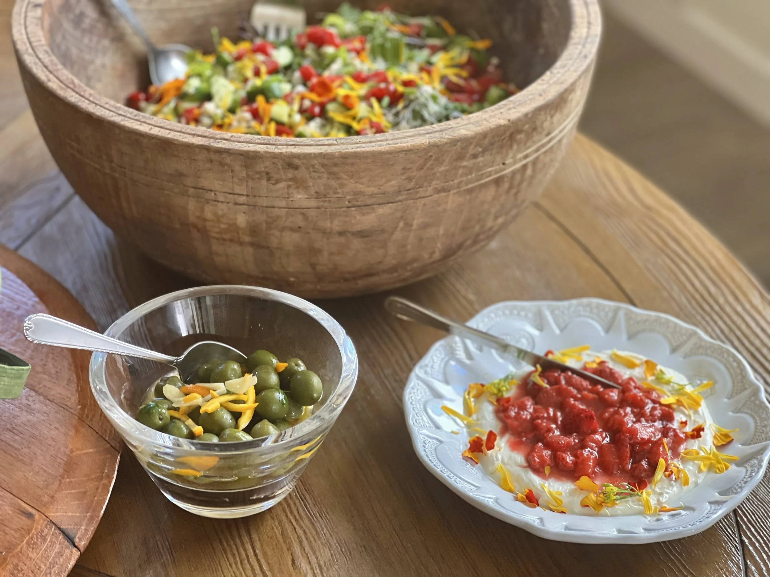 A bowl of colorful chopped salad, a small glass bowl of green olives with shredded cheese, and a plate of white yogurt topped with red sauce and edible flower petals on a wooden table.