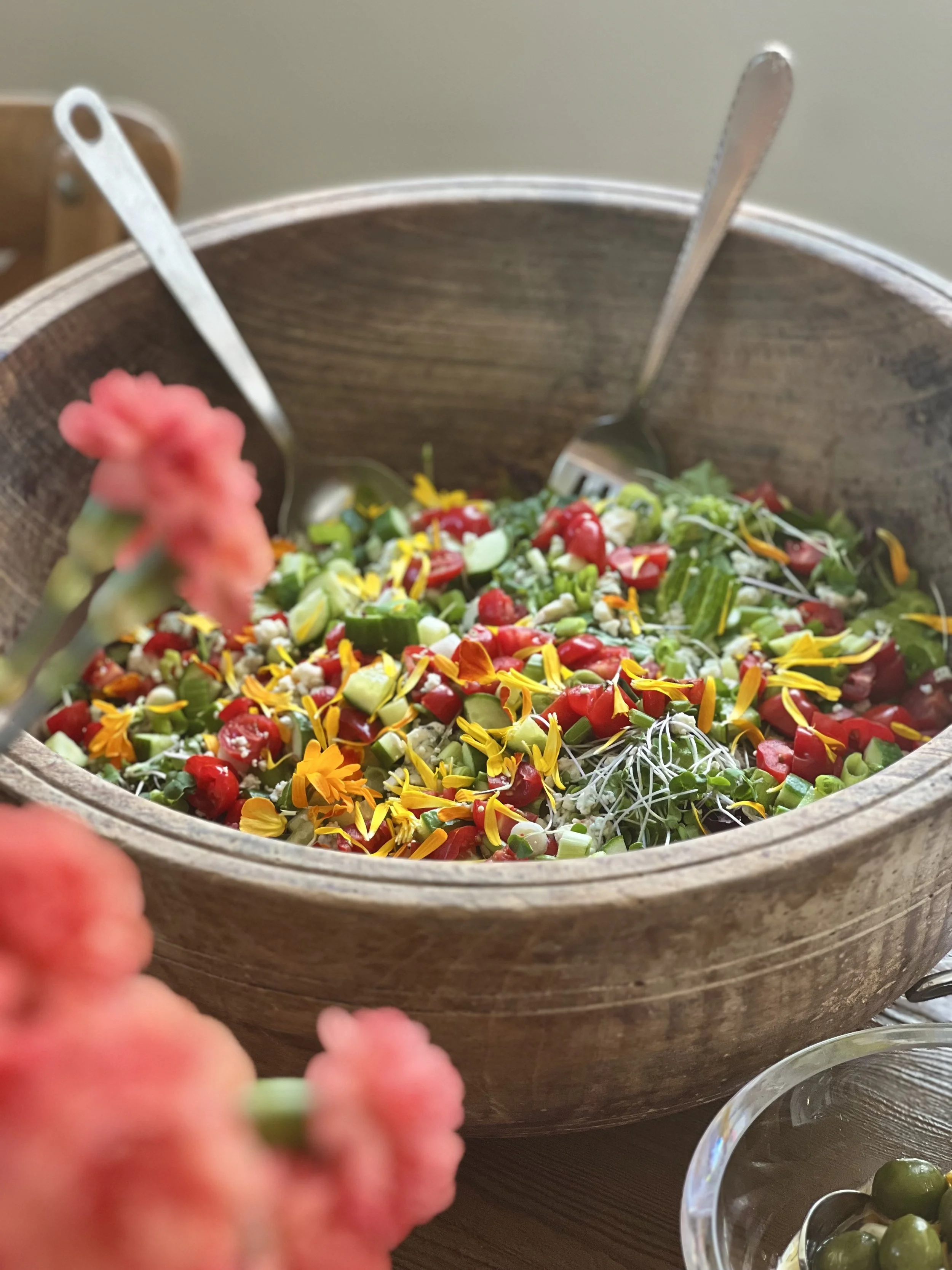 A large wooden bowl containing a colorful chopped vegetable salad with cherry tomatoes, bell peppers, onions, and edible flower petals, with a metal salad serving fork inside. Part of a smaller clear glass bowl with green olives is visible in the bottom right corner.