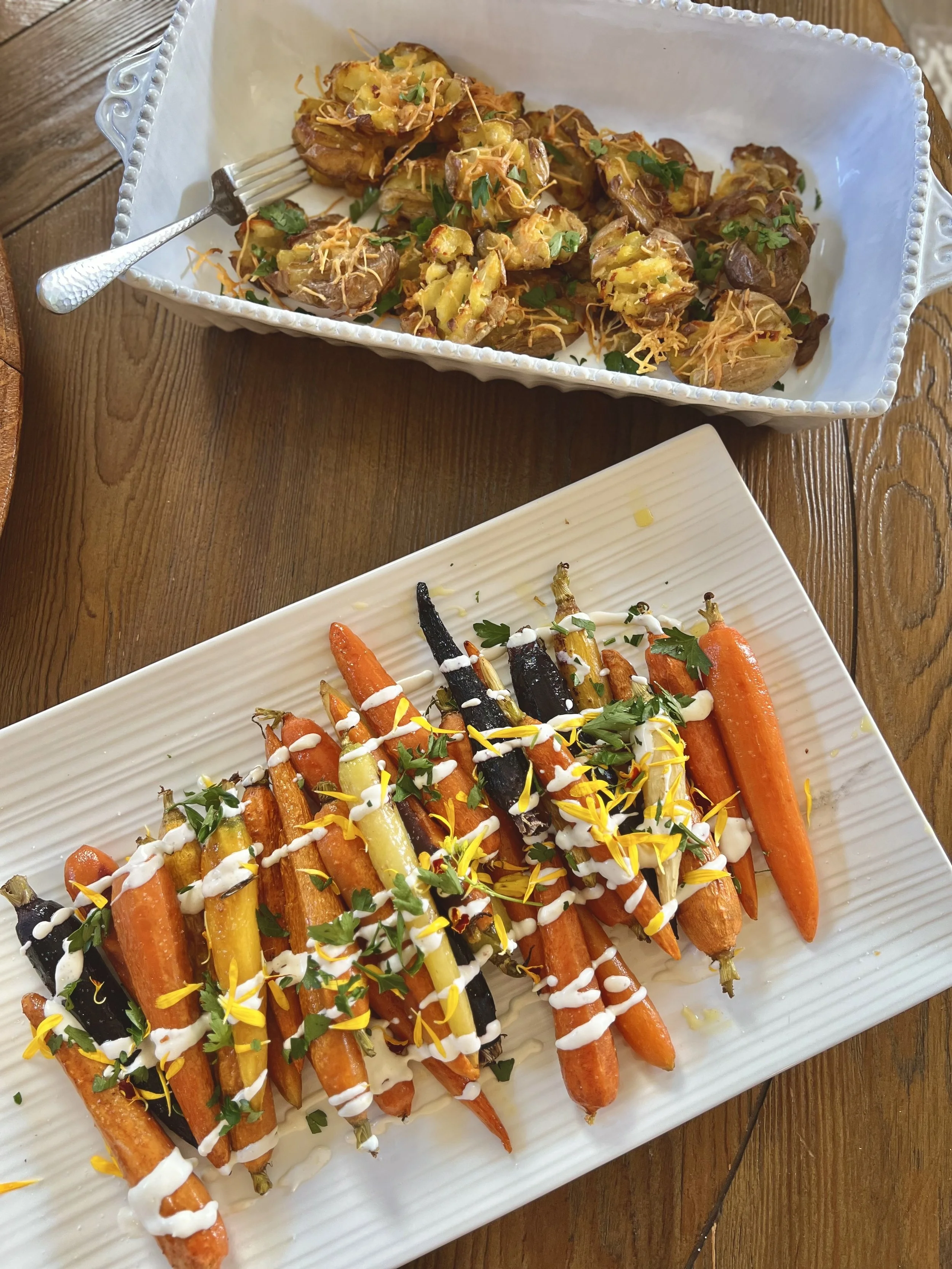 Roasted baby carrots on a rectangular white plate, drizzled with white sauce and garnished with herbs and edible flower petals. In a white decorative dish above, roasted potatoes topped with cheese, herbs, and shredded carrots.