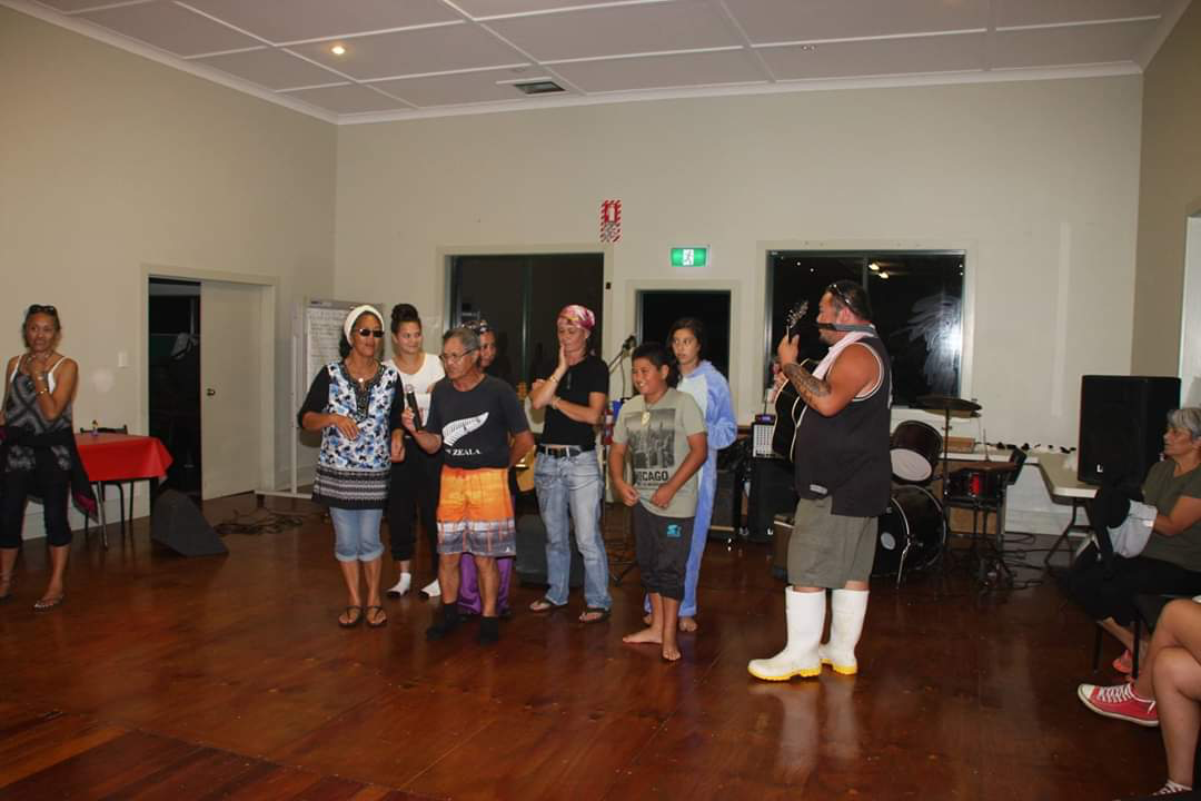Group of people in a room with musical instruments, one person playing guitar, others standing and clapping.