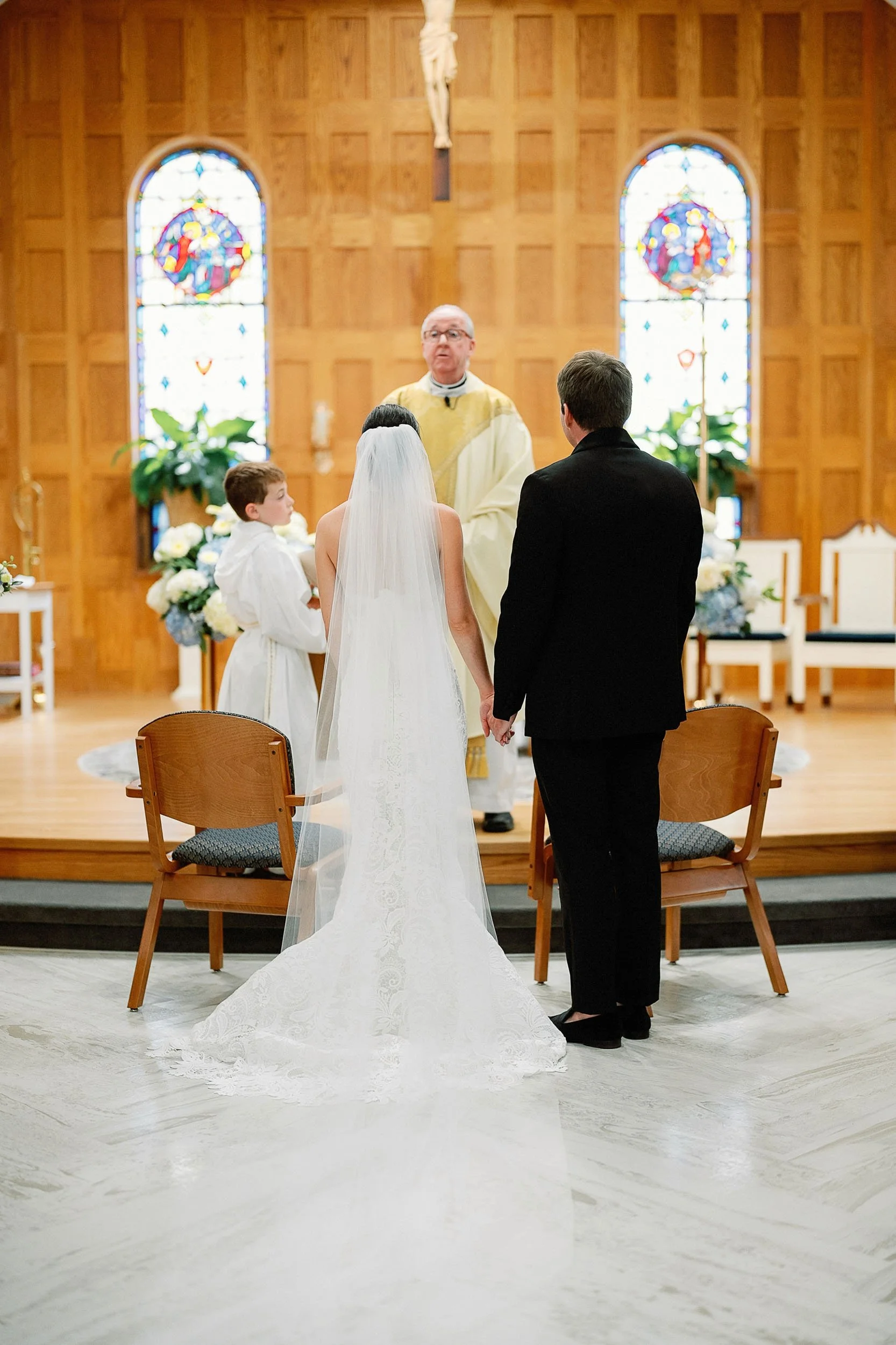 A wedding ceremony in a church with a bride and groom holding hands in front of a priest, with a young boy and a priest present, wooden altar with stained glass windows in the background.